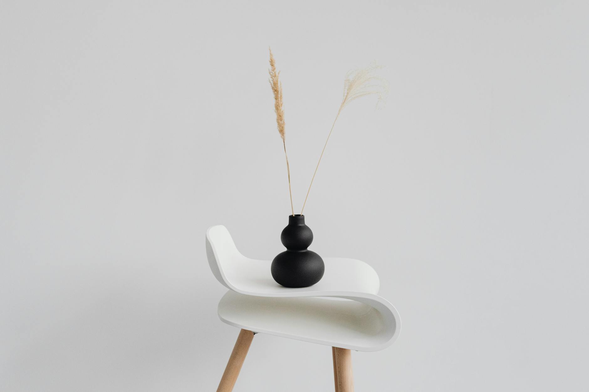 A sleek black vase with wheat ears on a modern white chair against a minimal background.