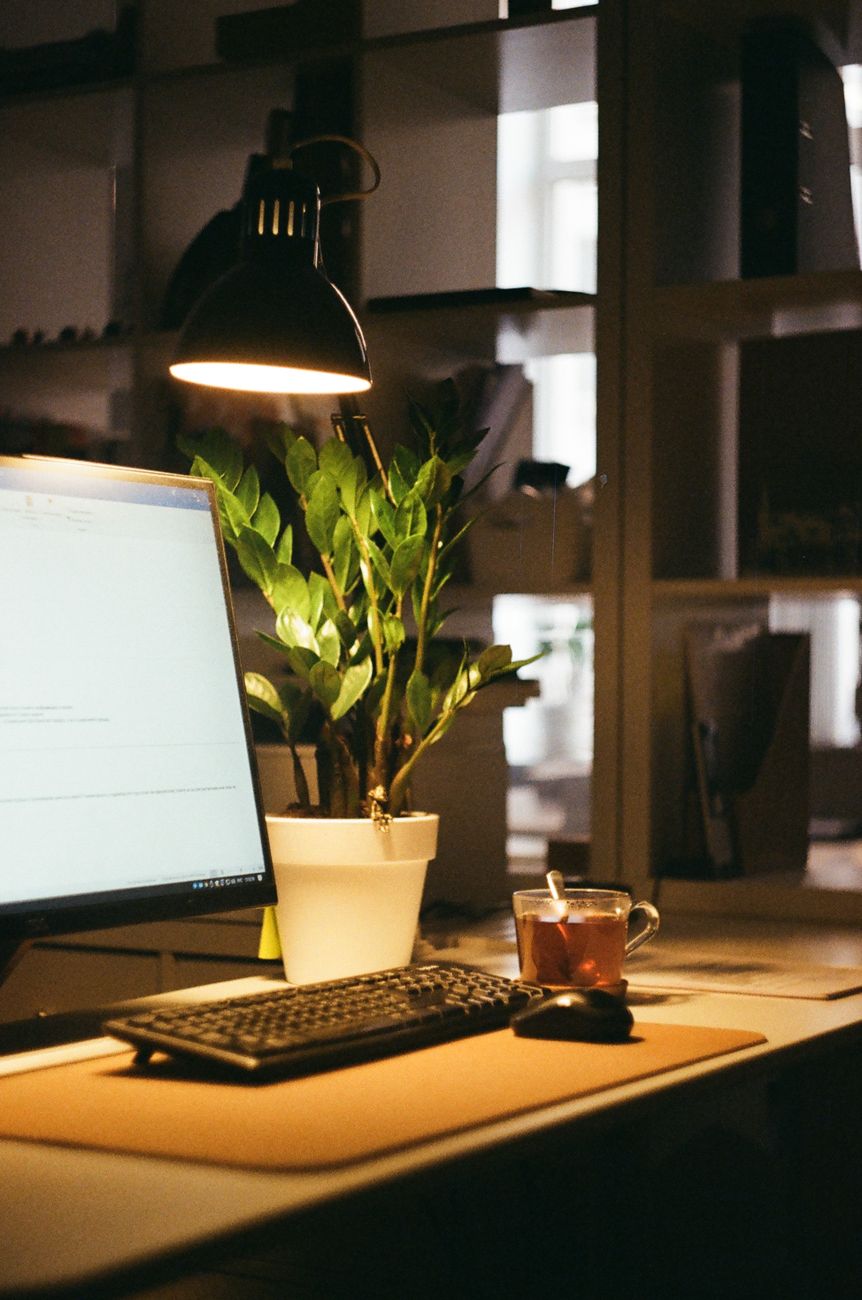 A warm-lit desk with a computer, lamp, plant, and tea cup in an office setup.