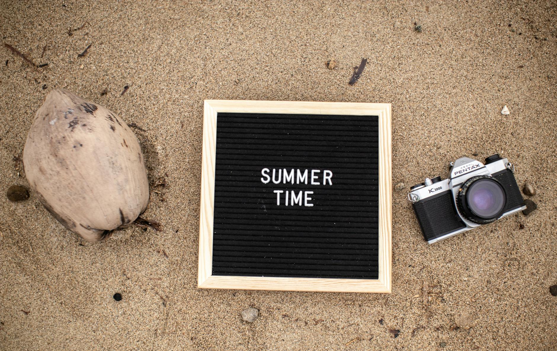 Letter board with SUMMER TIME text, vintage camera, and coconut on sandy beach.