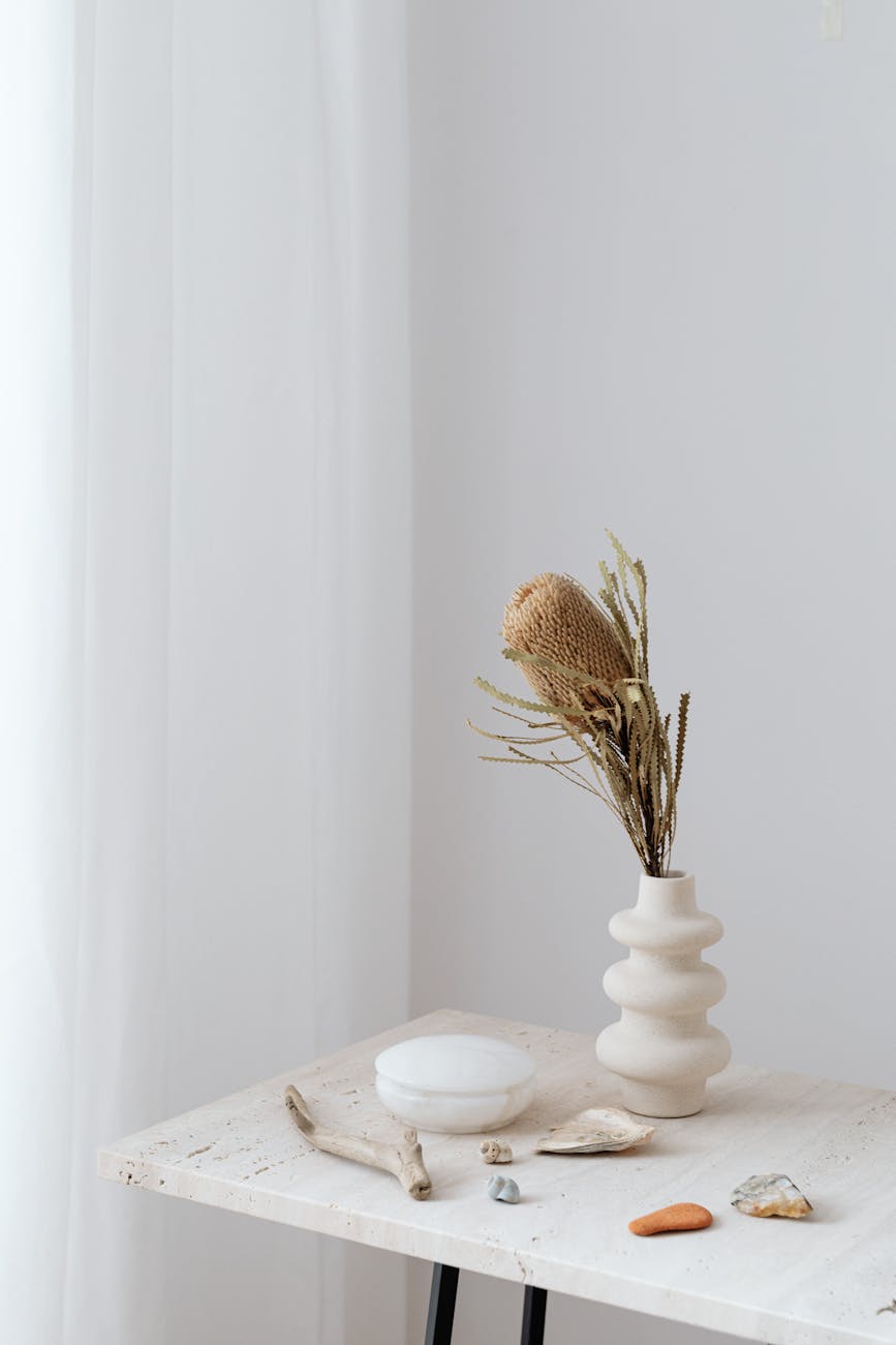 A serene still life of a table with a vase, stones, and driftwood against a white background.
