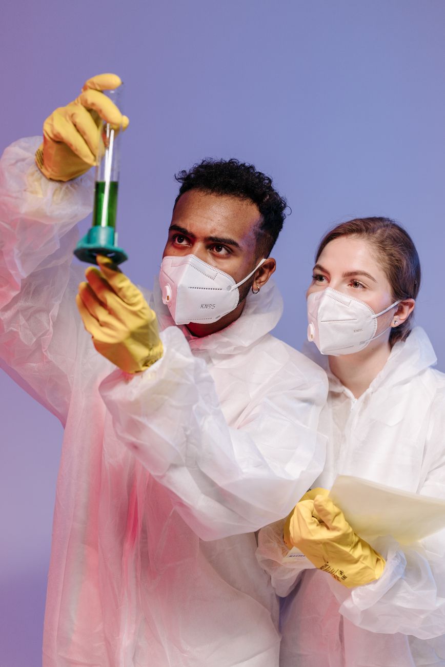 Two scientists in protective gear examine a green liquid in a laboratory setting.