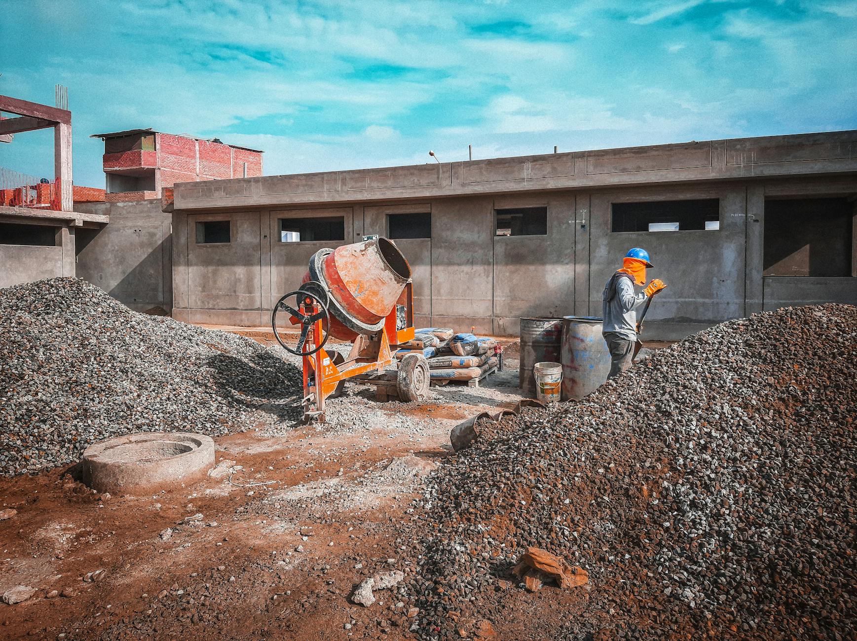 A construction site with a worker wearing safety gear next to a cement mixer and gravel piles.