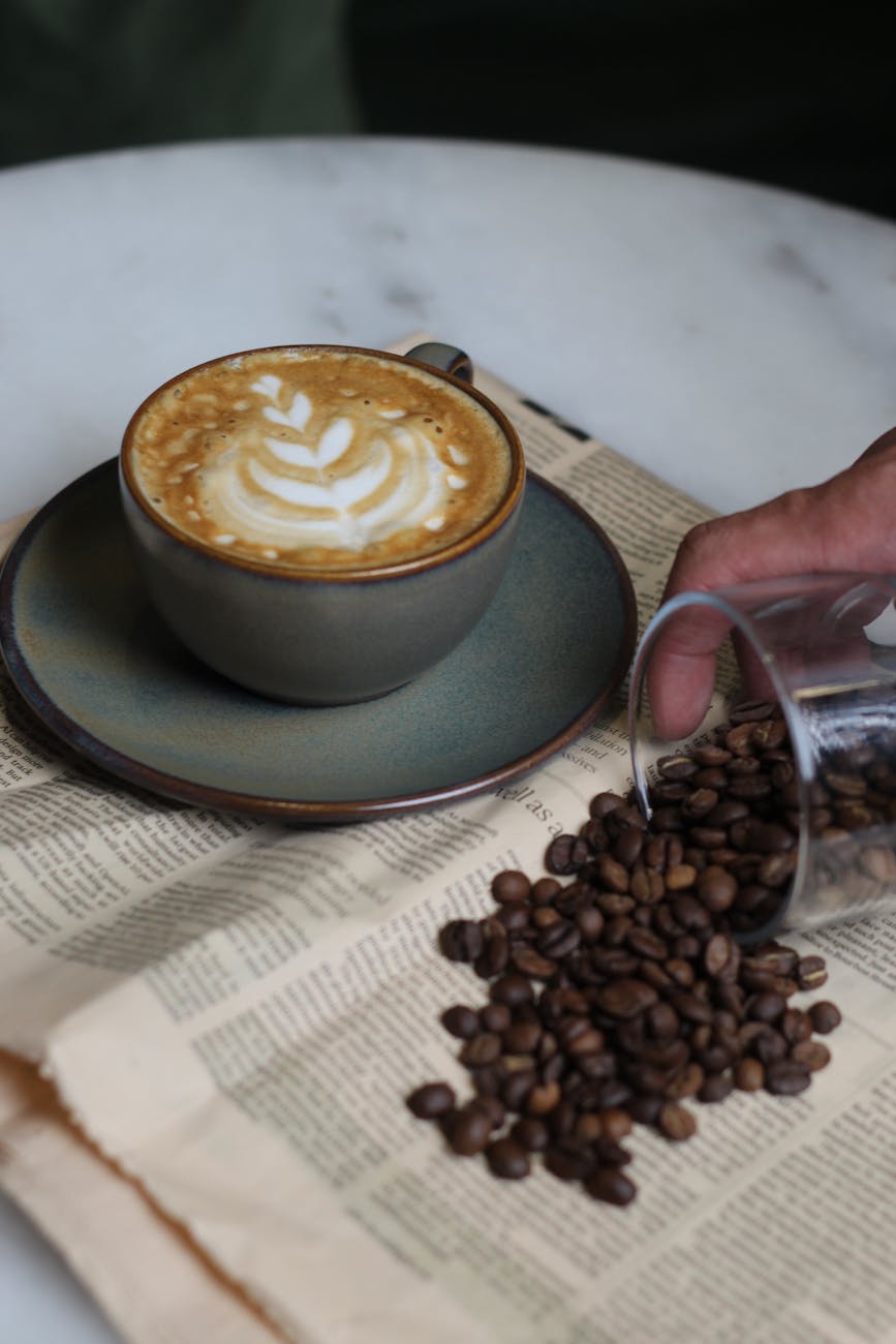 A cappuccino with latte art next to spilled coffee beans on a vintage newspaper.