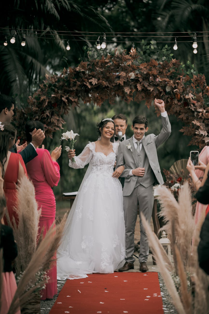 Happy couple celebrates their wedding under a floral arch with loved ones cheering on a festive day.