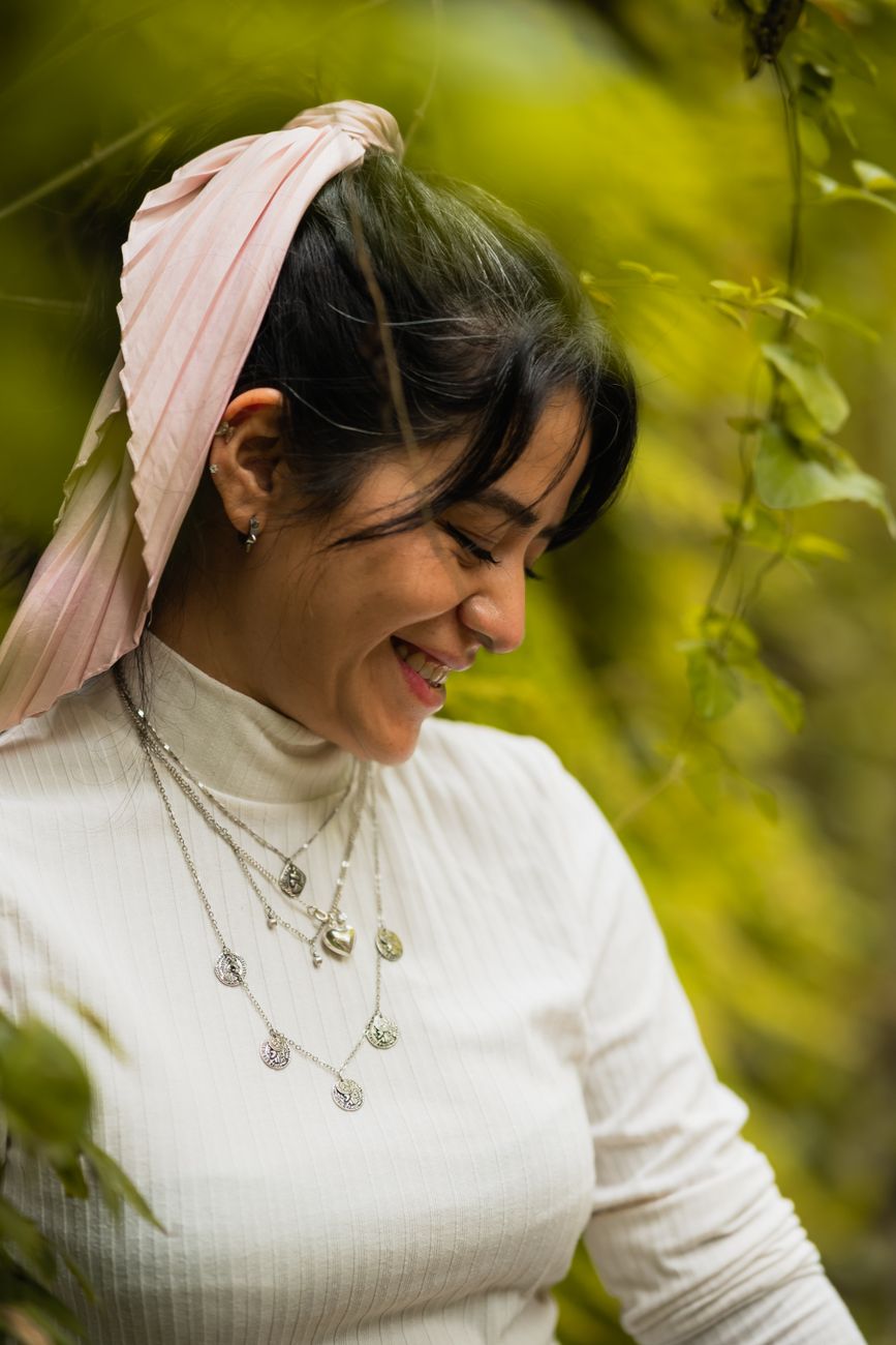 A young woman with a pink scarf smiles amidst lush greenery, showcasing layered necklaces outdoors.