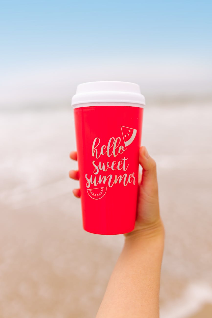 Bright red cup with 'Hello Sweet Summer' text held on a beach. Perfect for summer vibes.