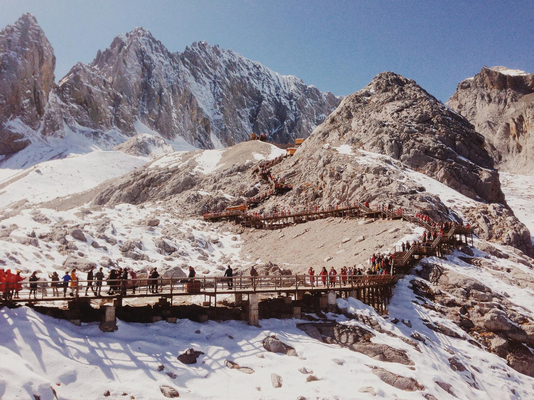 Tourists hiking wooden bridges amidst snowcapped peaks in Lijiang, China.