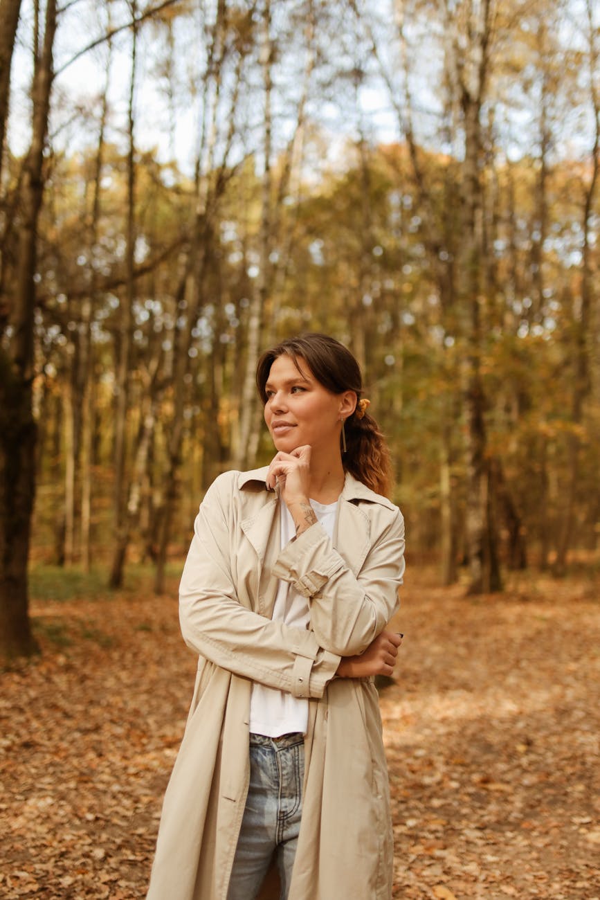 Woman in a trench coat standing thoughtfully in an autumn forest scene.