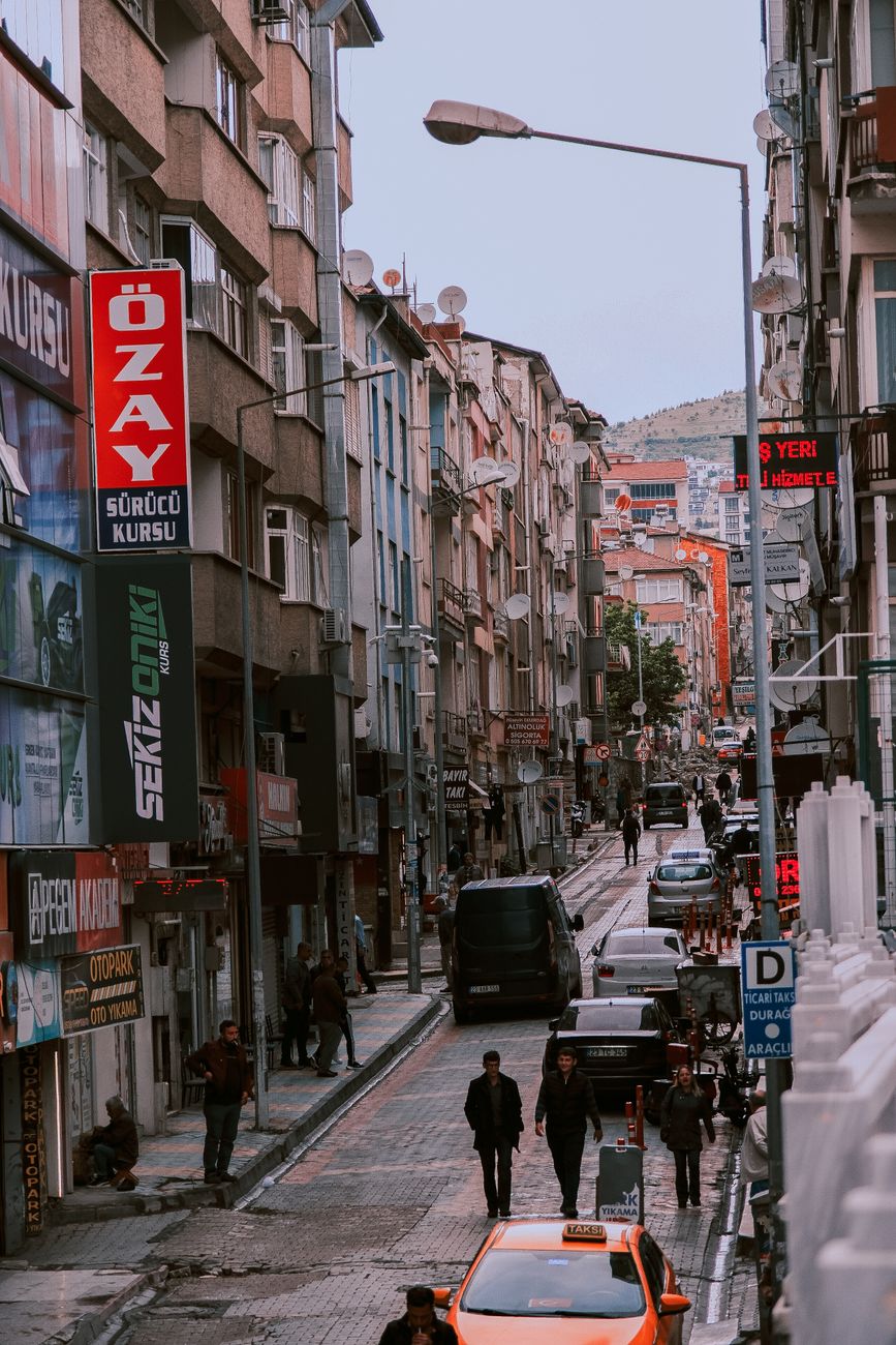 Vibrant city street scene with people walking and cars on a busy urban road, lined with buildings.
