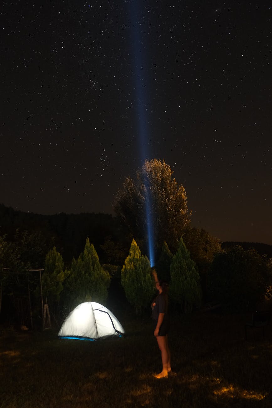 A woman standing by a tent at night, flashlight beam pointing at a starry sky.