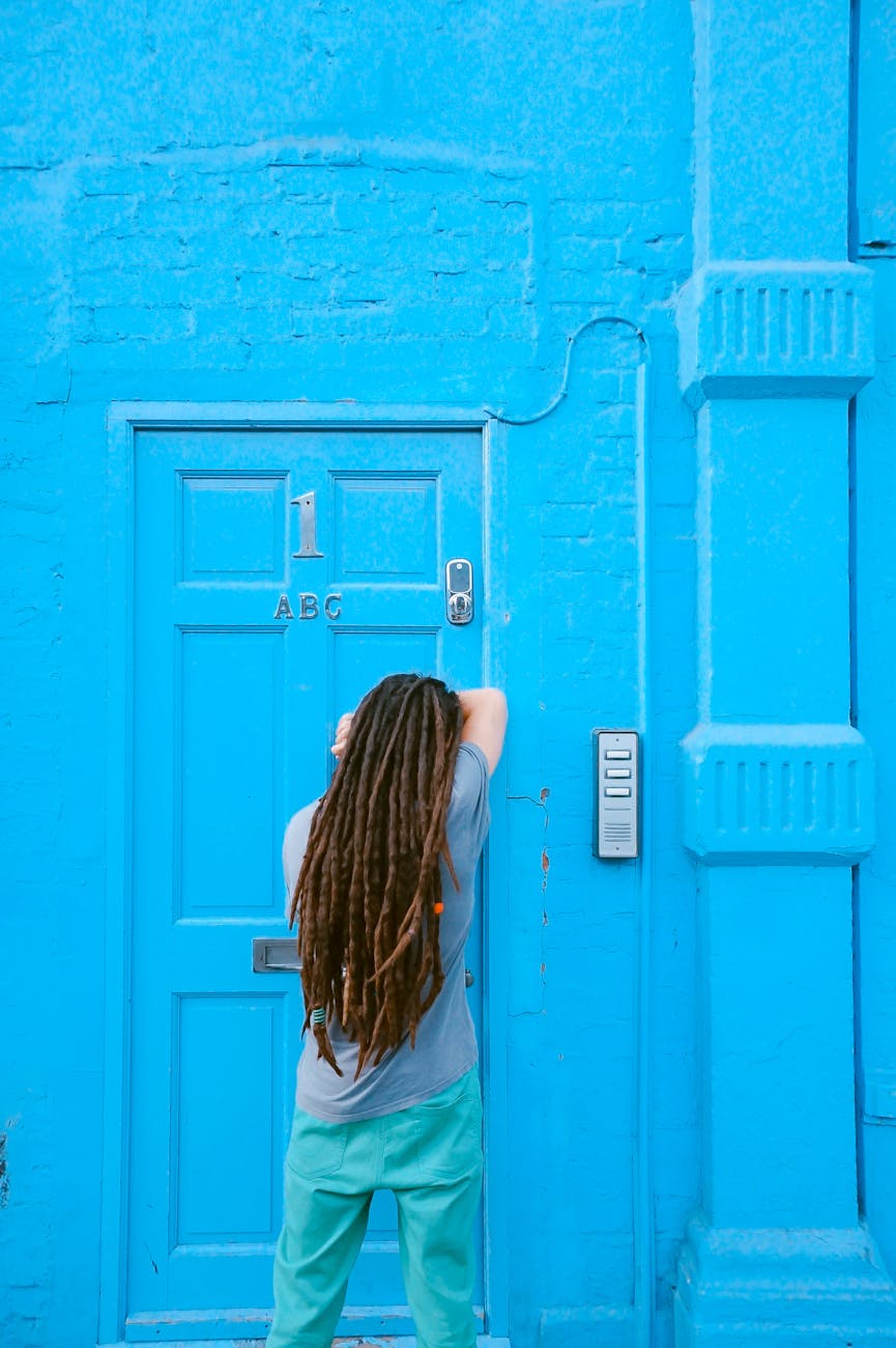 A man with long hair stands outside a bright blue door in an urban setting.