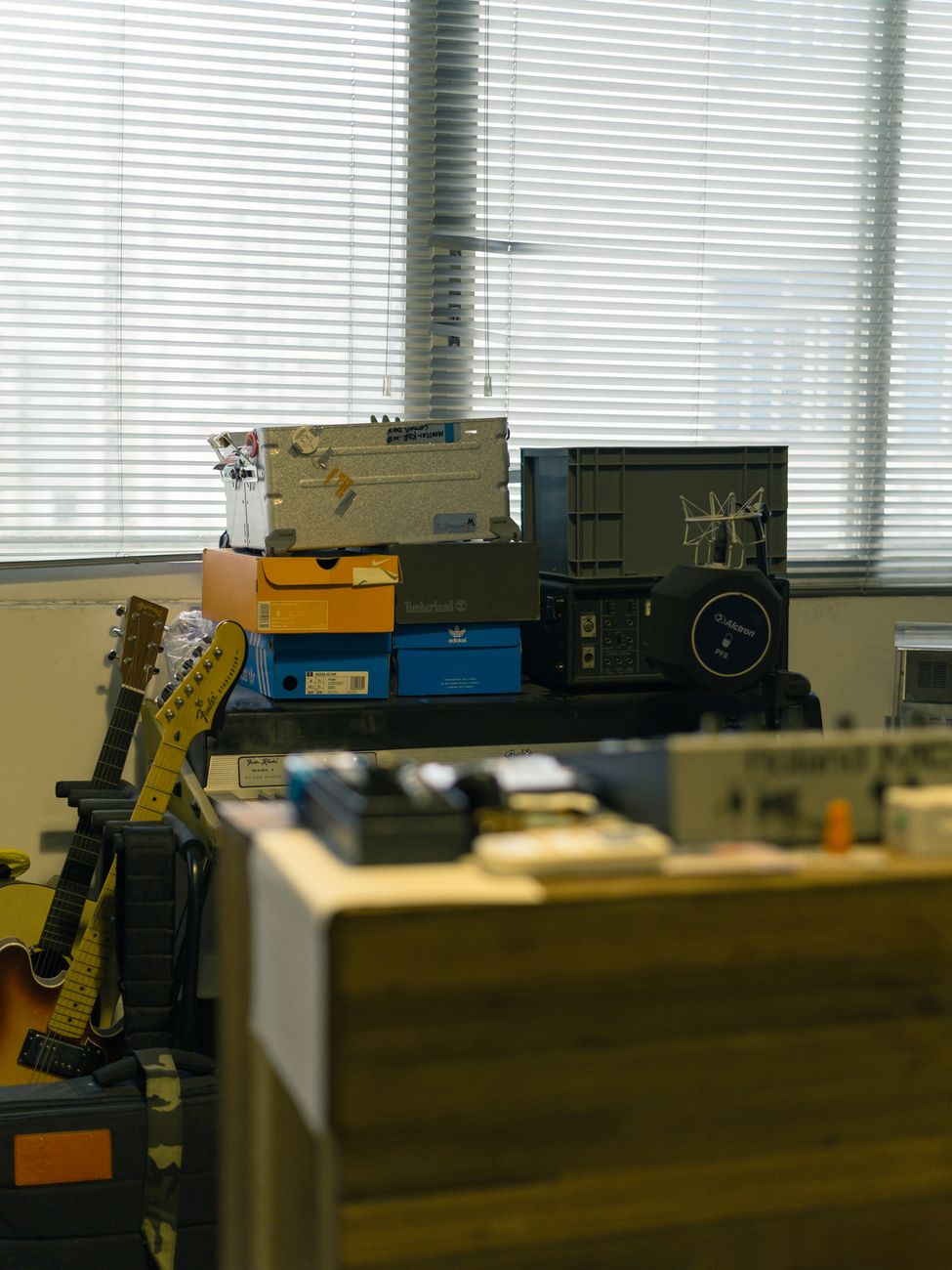 A cluttered office corner with stacked boxes and guitars against a window.