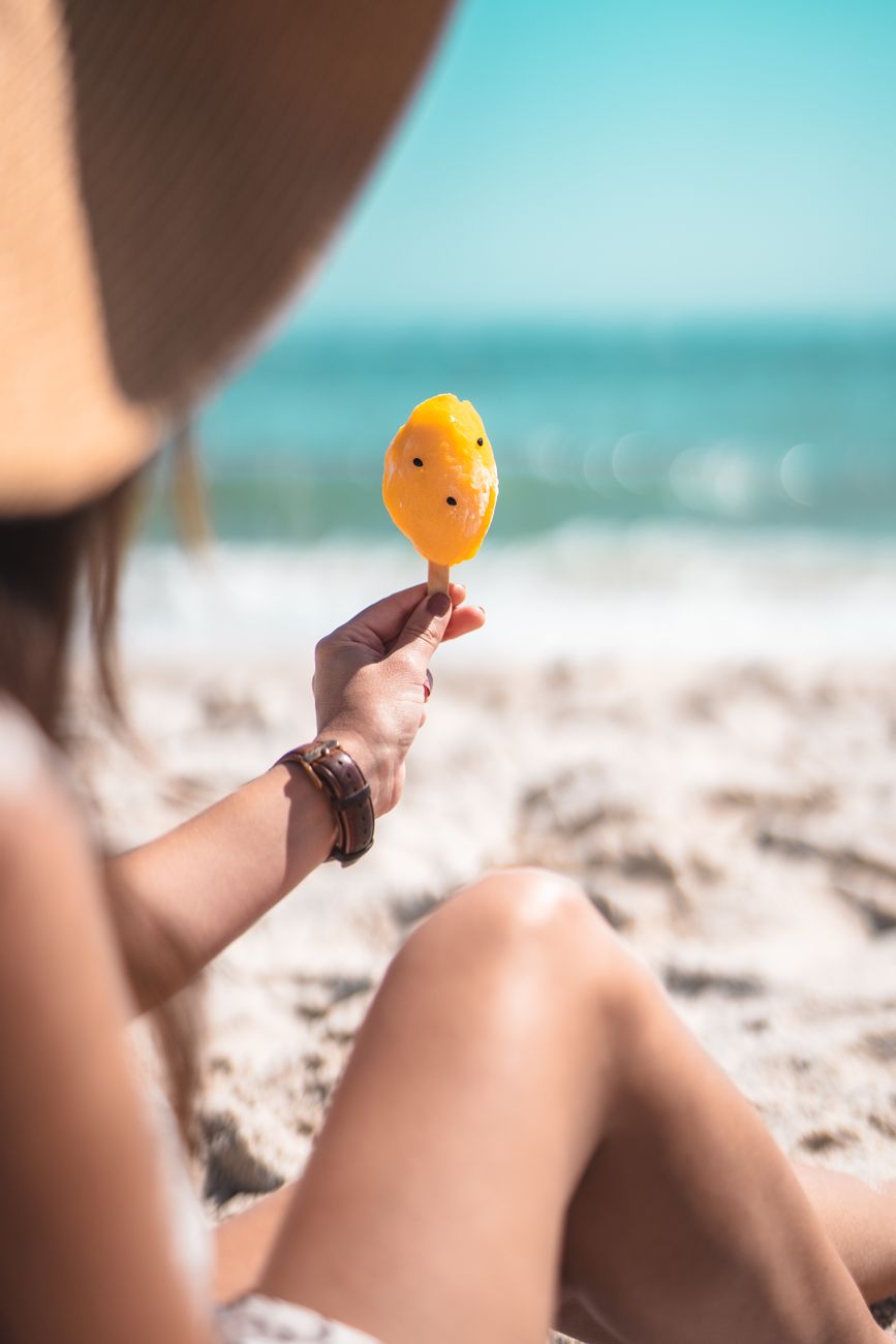 A woman enjoys a sunny summer day at the beach holding a refreshing orange popsicle.