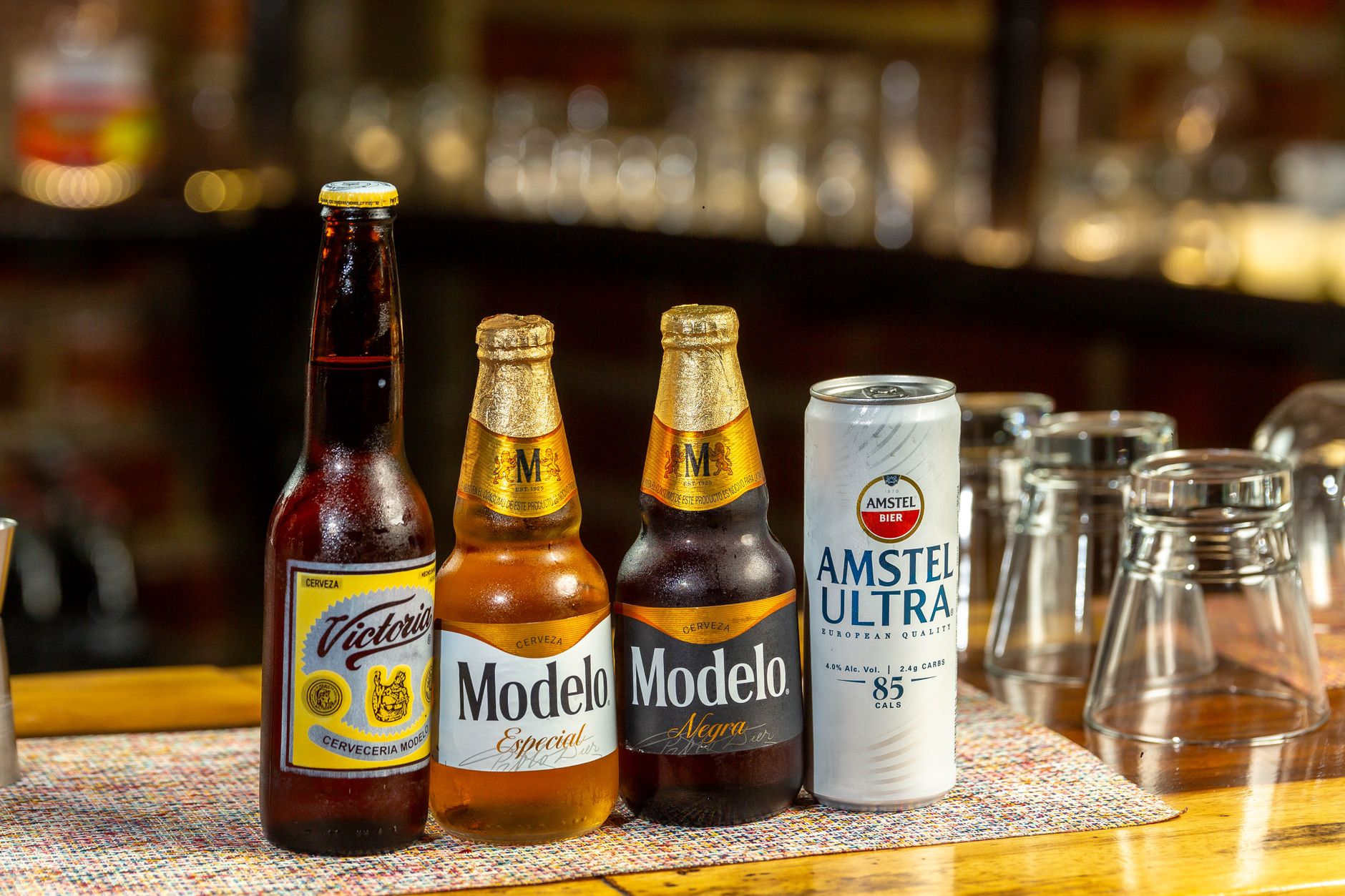 A selection of beer bottles and a can on a bar counter, featuring popular brands like Victoria, Modelo, and Amstel Ultra.