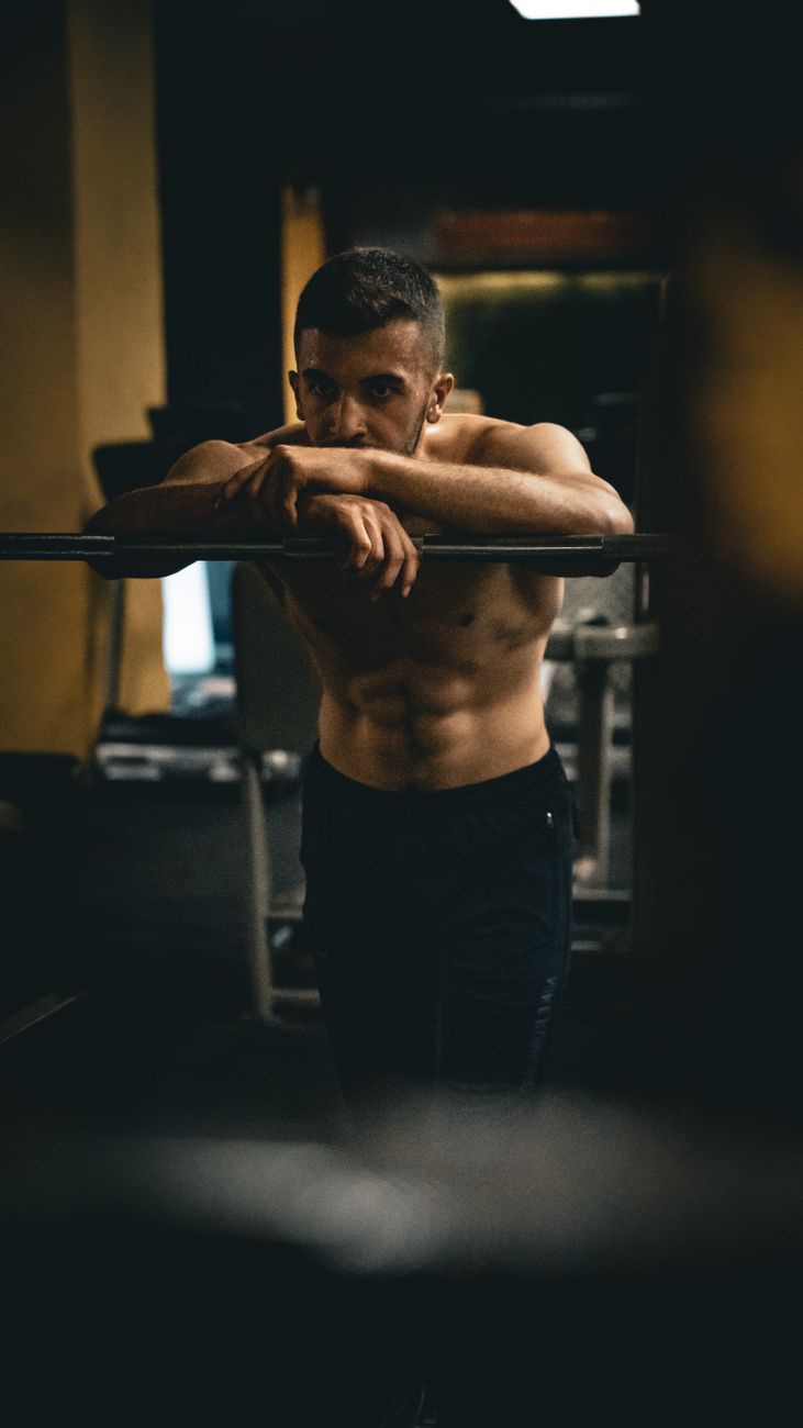 A topless man with short hair leaning on a barbell in a dimly lit gym, showcasing fitness and muscle.