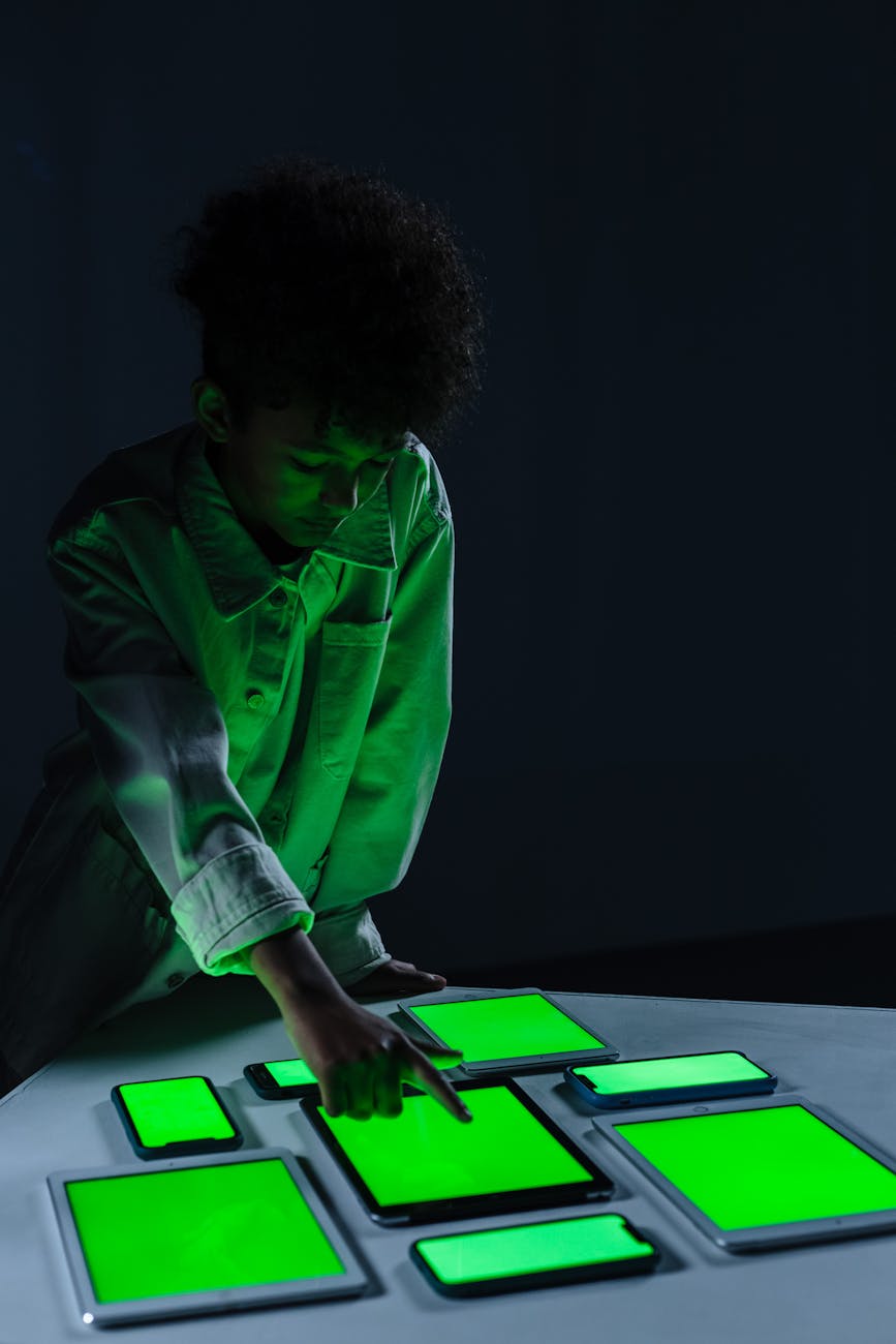 A child in a dark room interacts with several digital screens, glowing green, illustrating modern technology use.