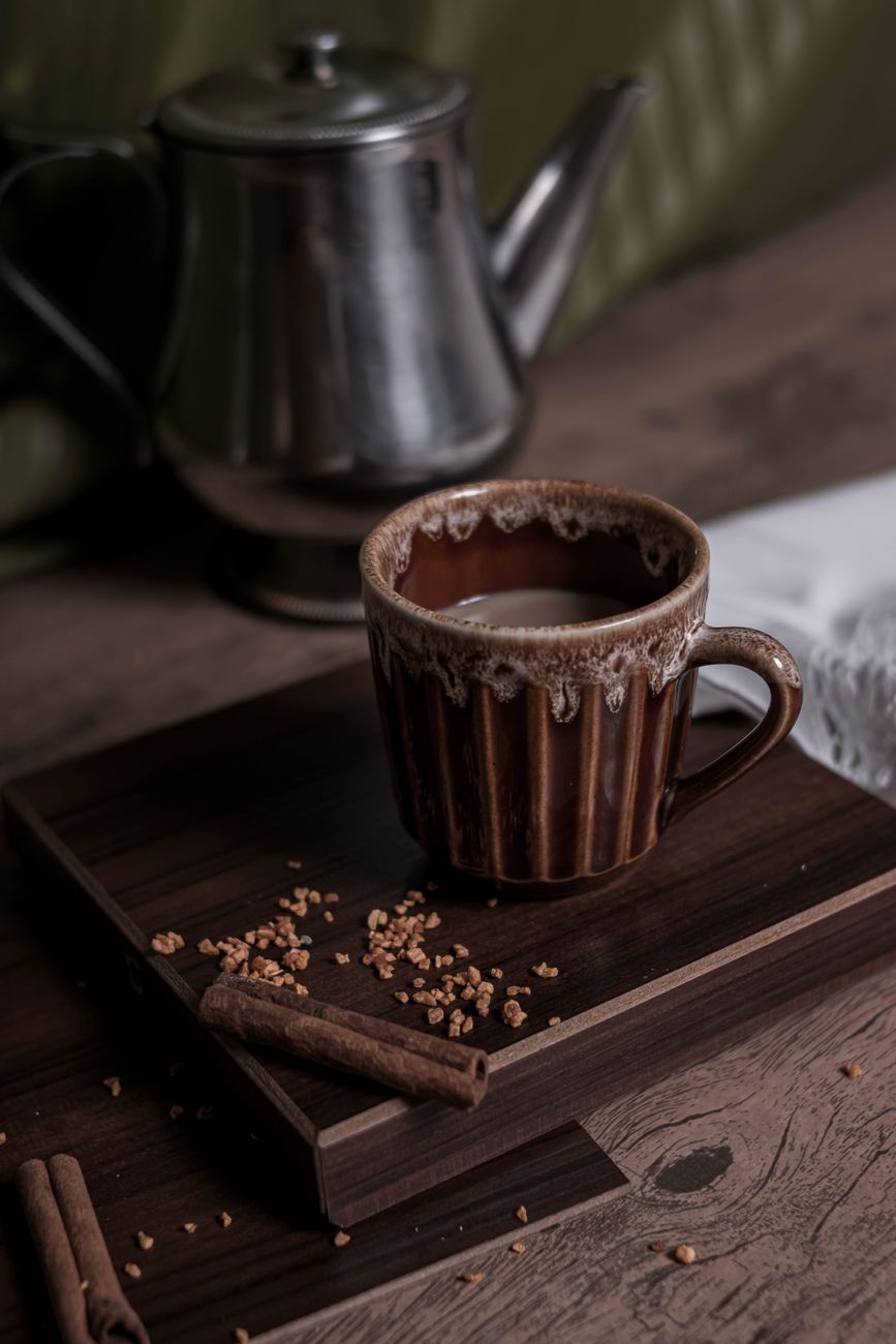 A rustic coffee setup with a mug and kettle, enhanced with cinnamon sticks and sugar crystals.