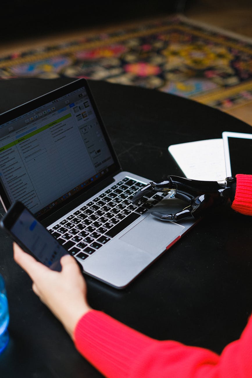 Close-up of a person with a prosthetic hand typing on a laptop while holding a phone, showcasing modern technology.