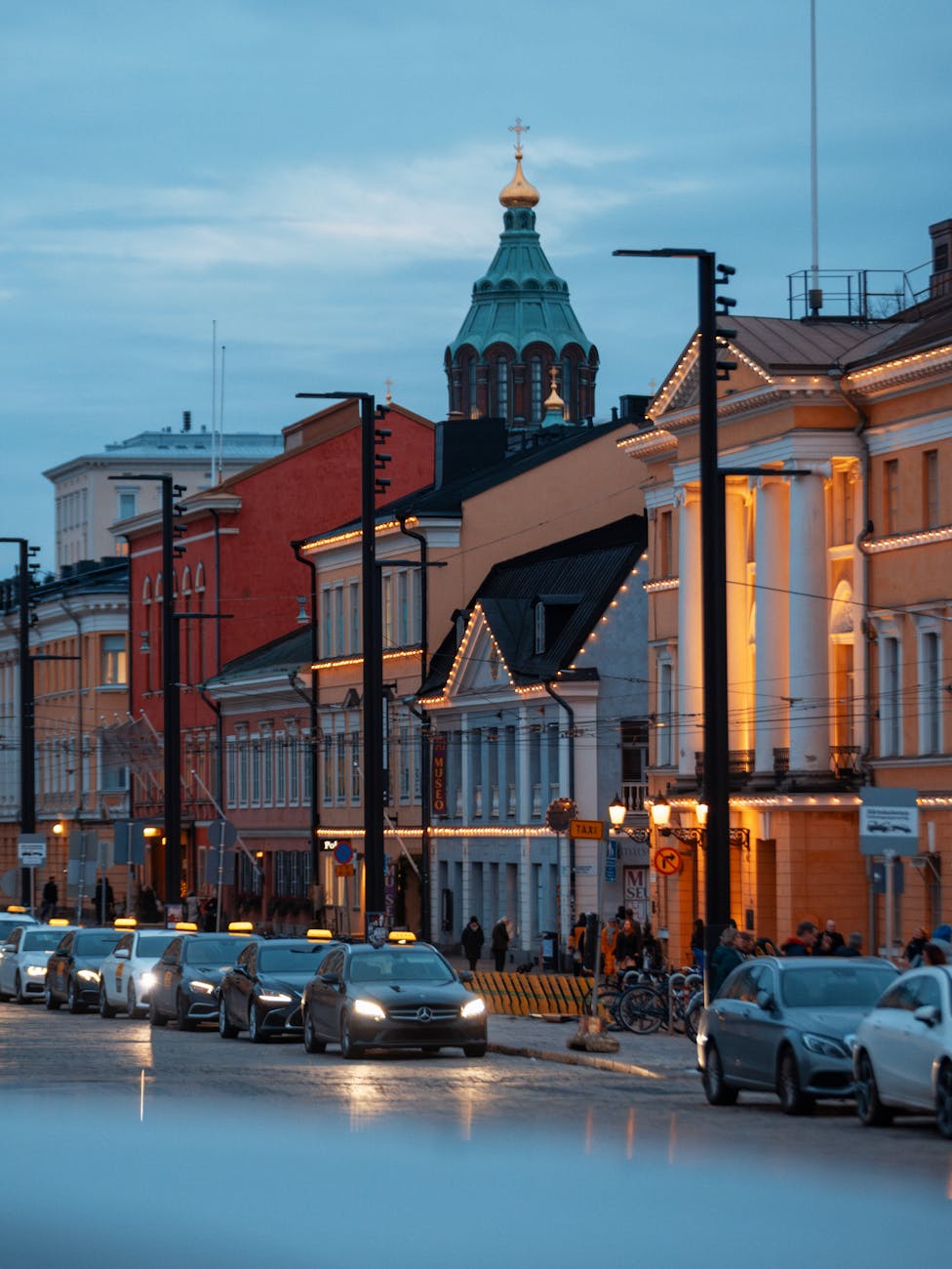 Twilight view of a historic street in Helsinki, Finland, featuring lit buildings and passing cars.