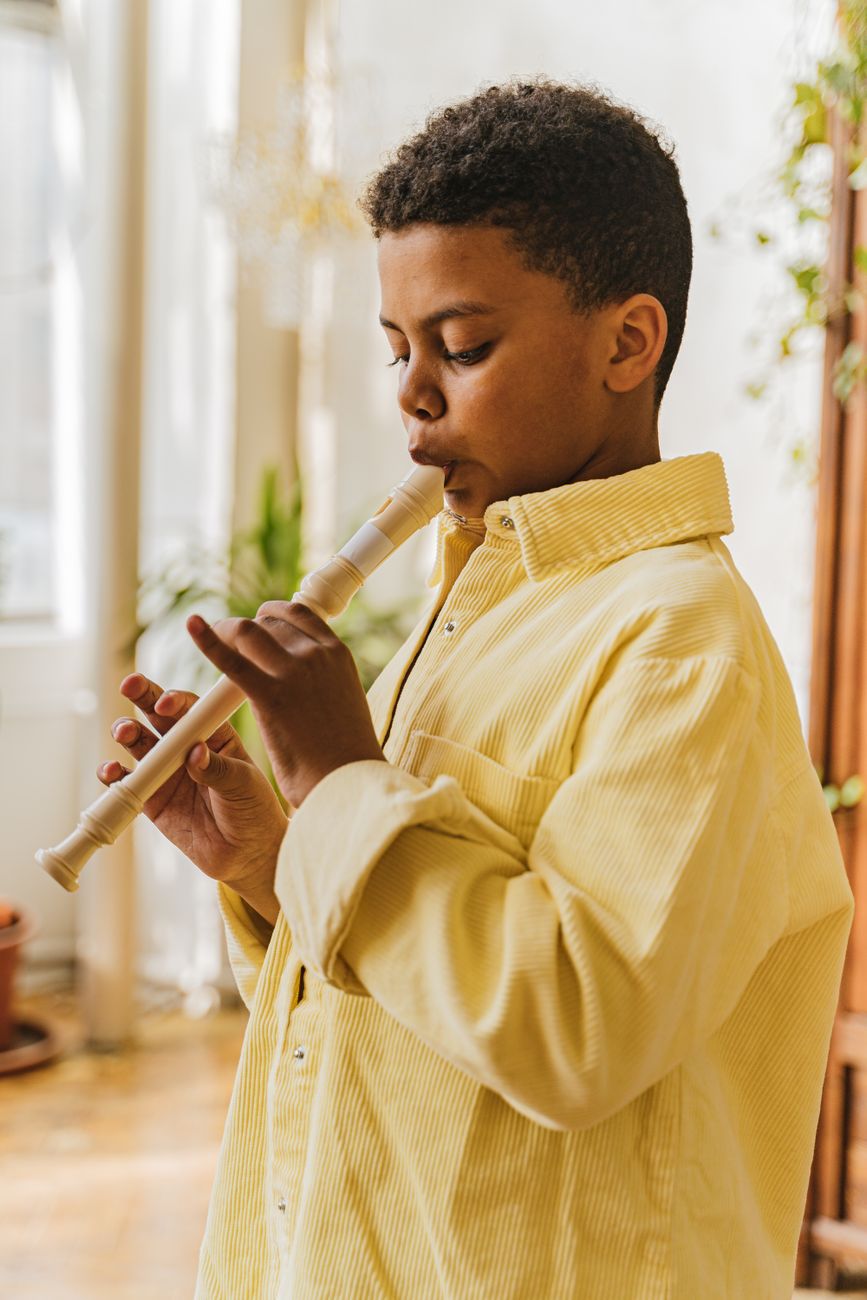 A young boy concentrates while playing the flute indoors, bathed in natural light.