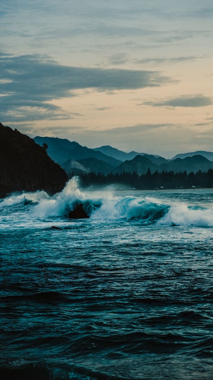 Majestic waves crash against Aceh's coastline, framed by distant mountain ranges under a moody sky.