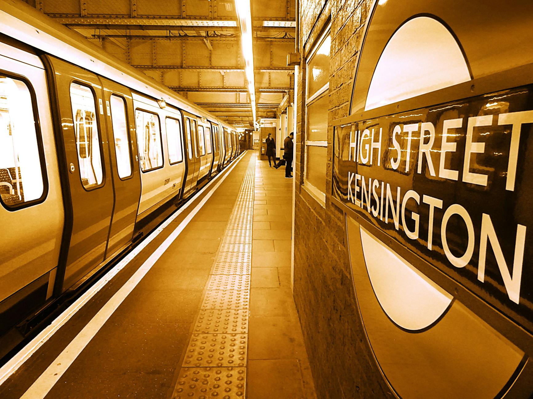A sepia-toned view of High Street Kensington station platform with a train and commuters in London.
