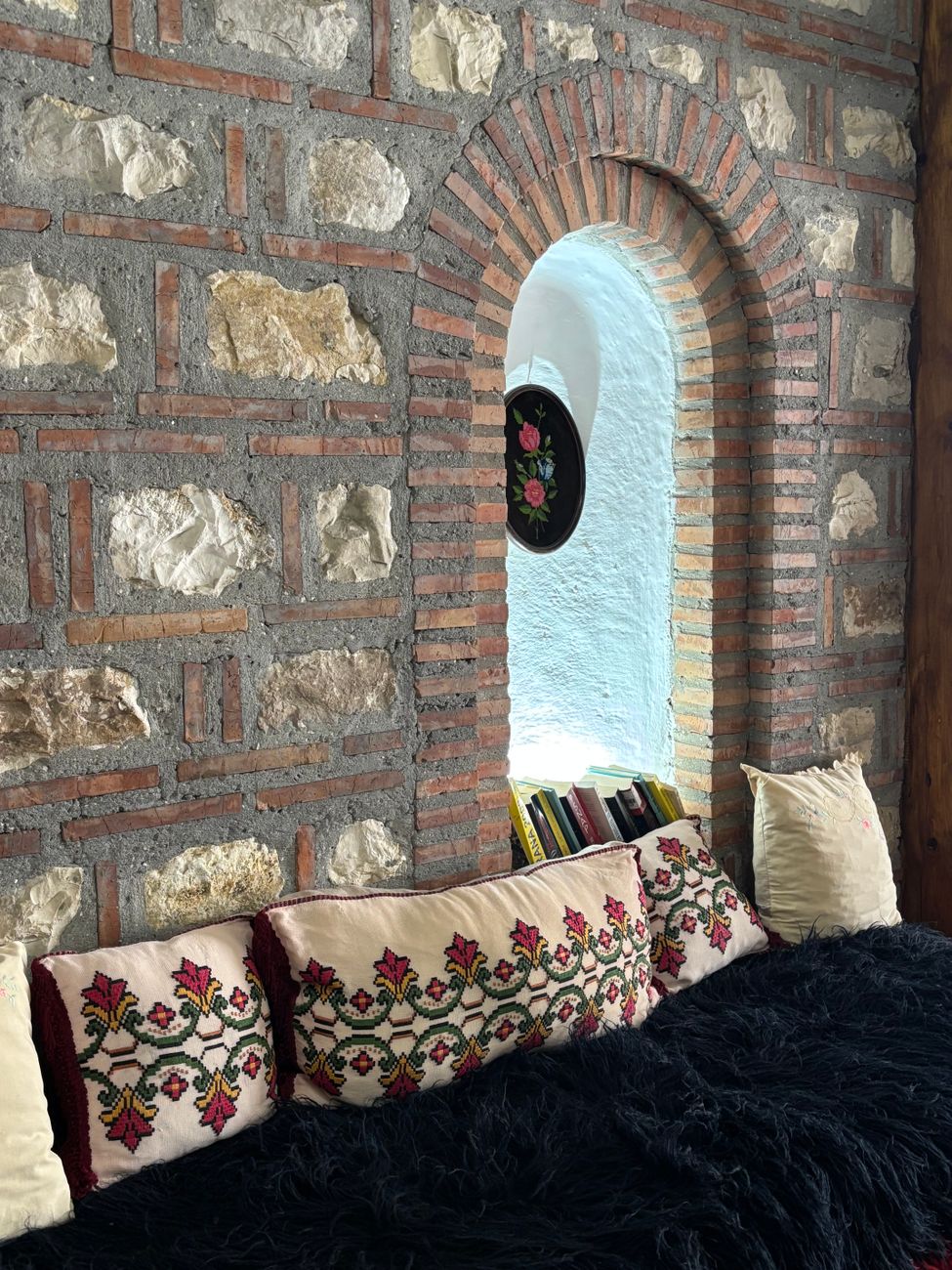 Inviting alcove with a stone wall, embroidered pillows, and an arched window, featuring a rustic interior design.