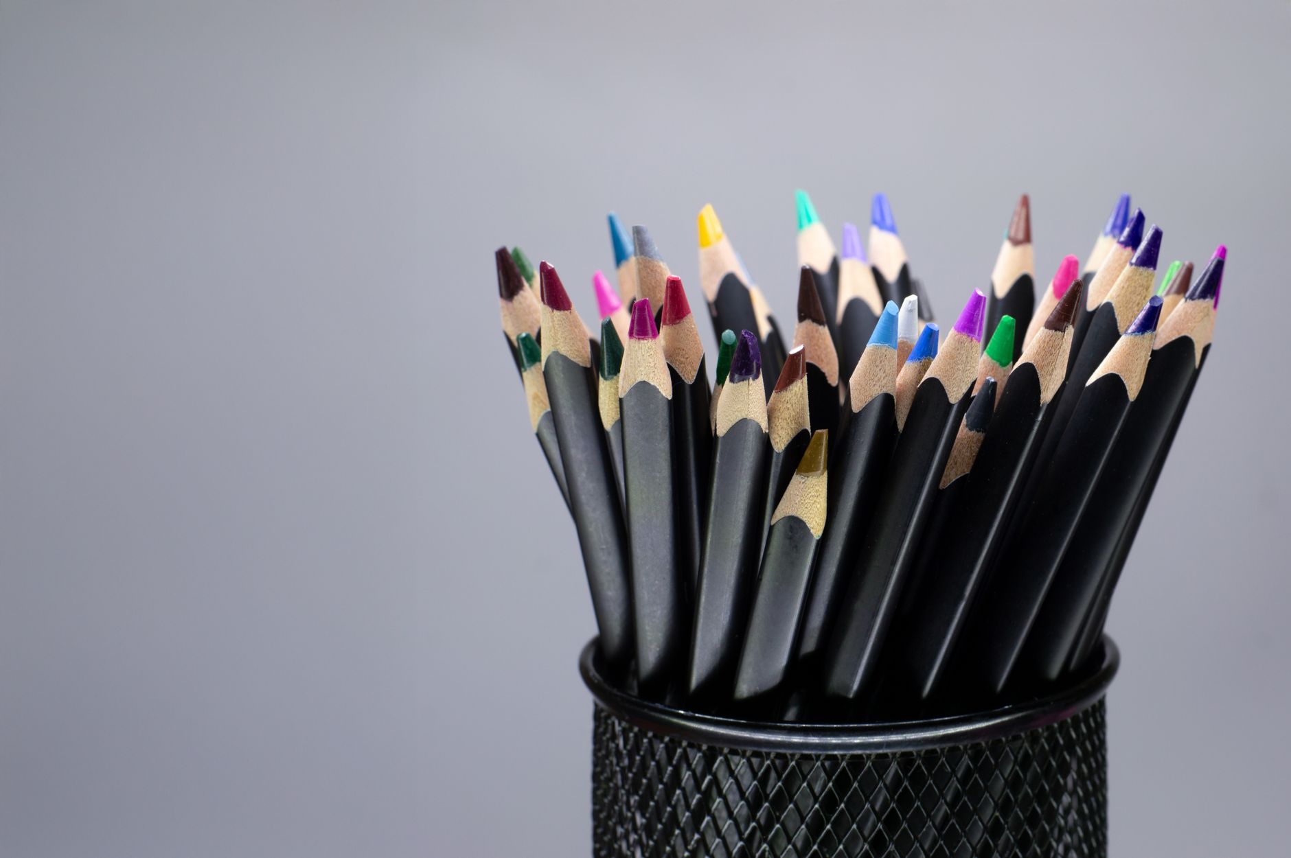 A collection of colorful pencils in a black mesh holder against a gray background.
