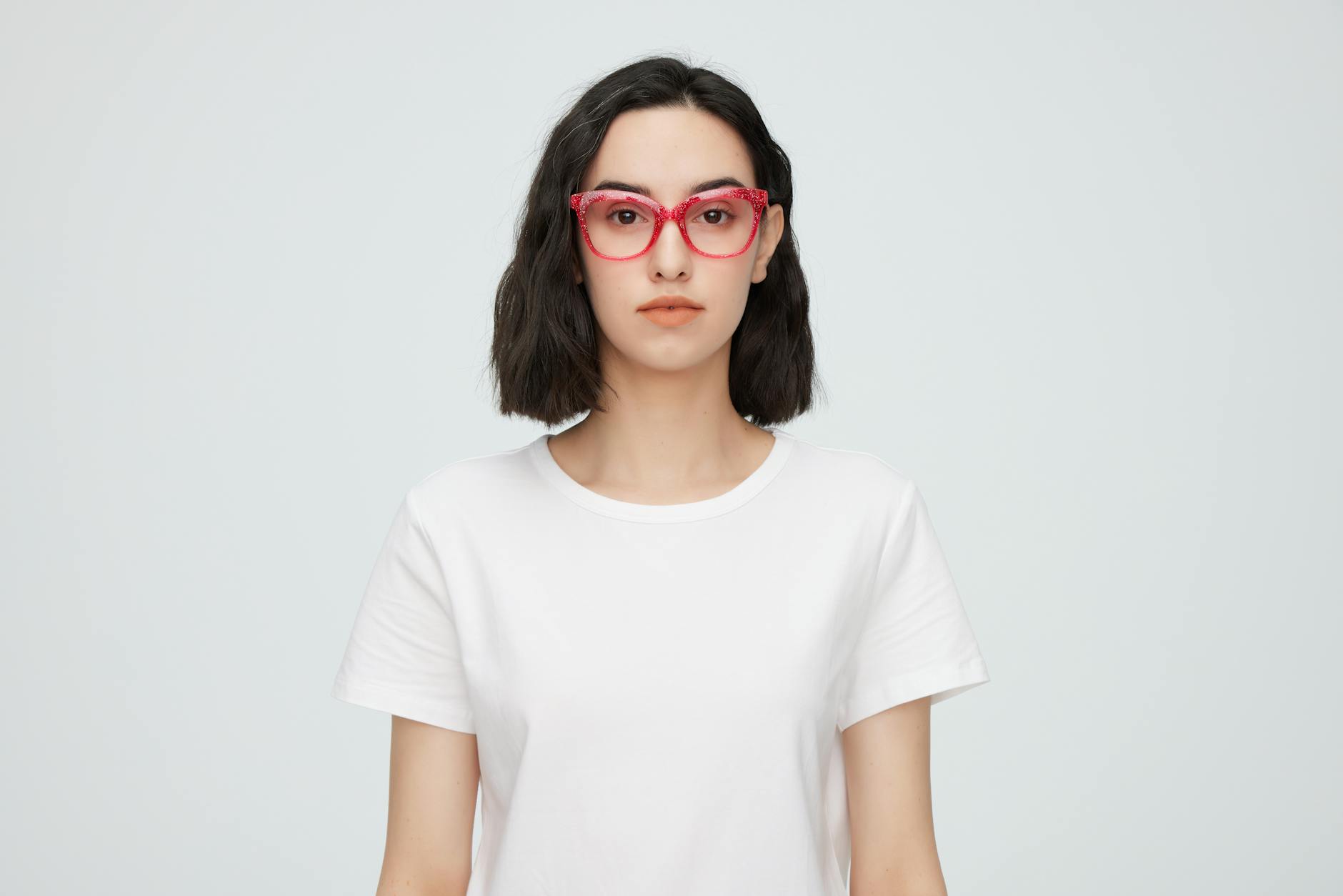 Portrait of a young woman wearing red eyeglasses and a white shirt against a plain background.