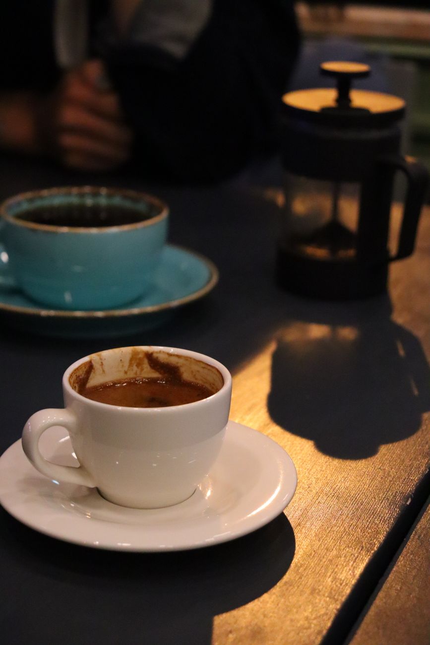 A close-up shot of a coffee cup on a saucer with a French press in the background on a wooden table.