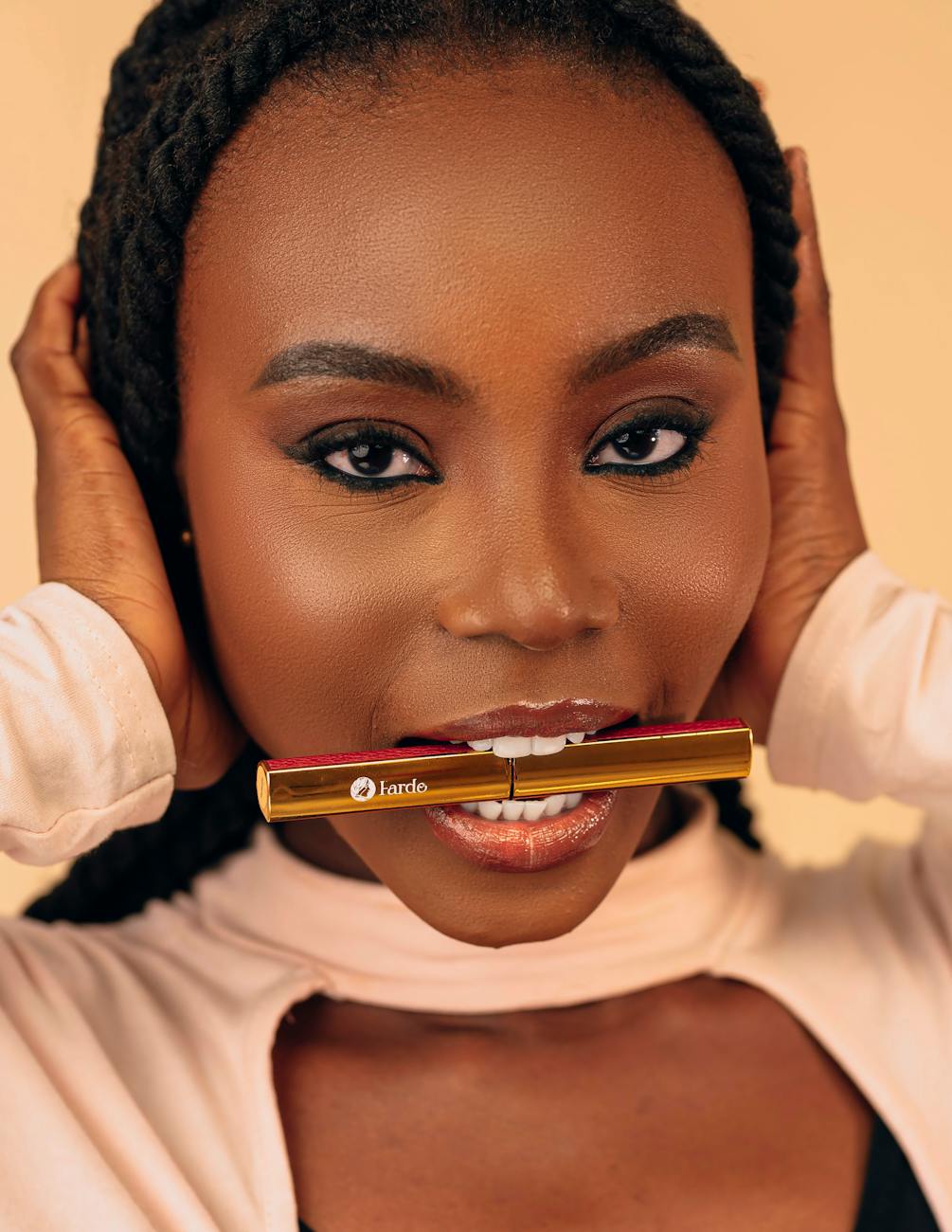 Close-up portrait of a woman with bold makeup holding a lipstick between her teeth.