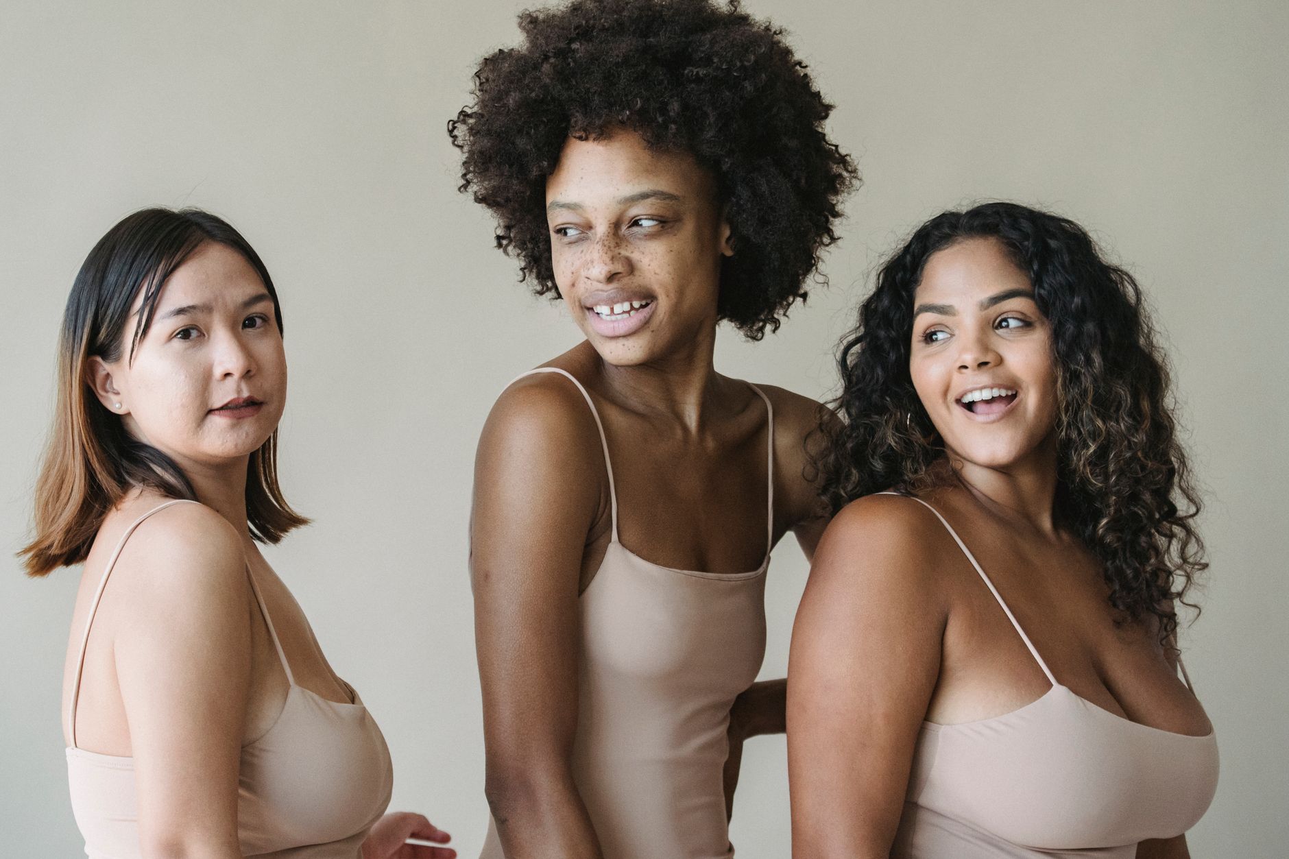 Three women of different ethnicities smiling and posing in neutral-toned swimwear.