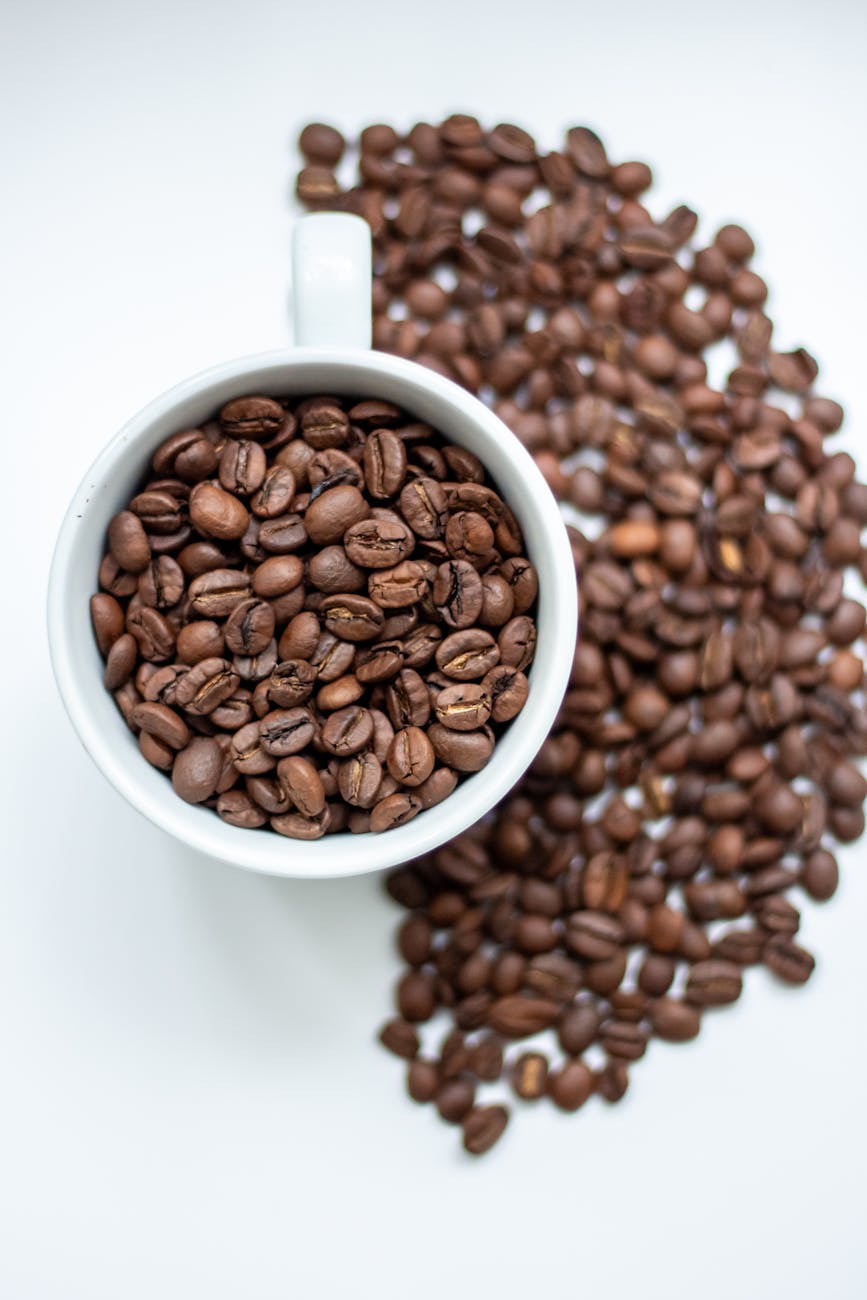 High-angle view of a white cup filled with coffee beans on a white surface.