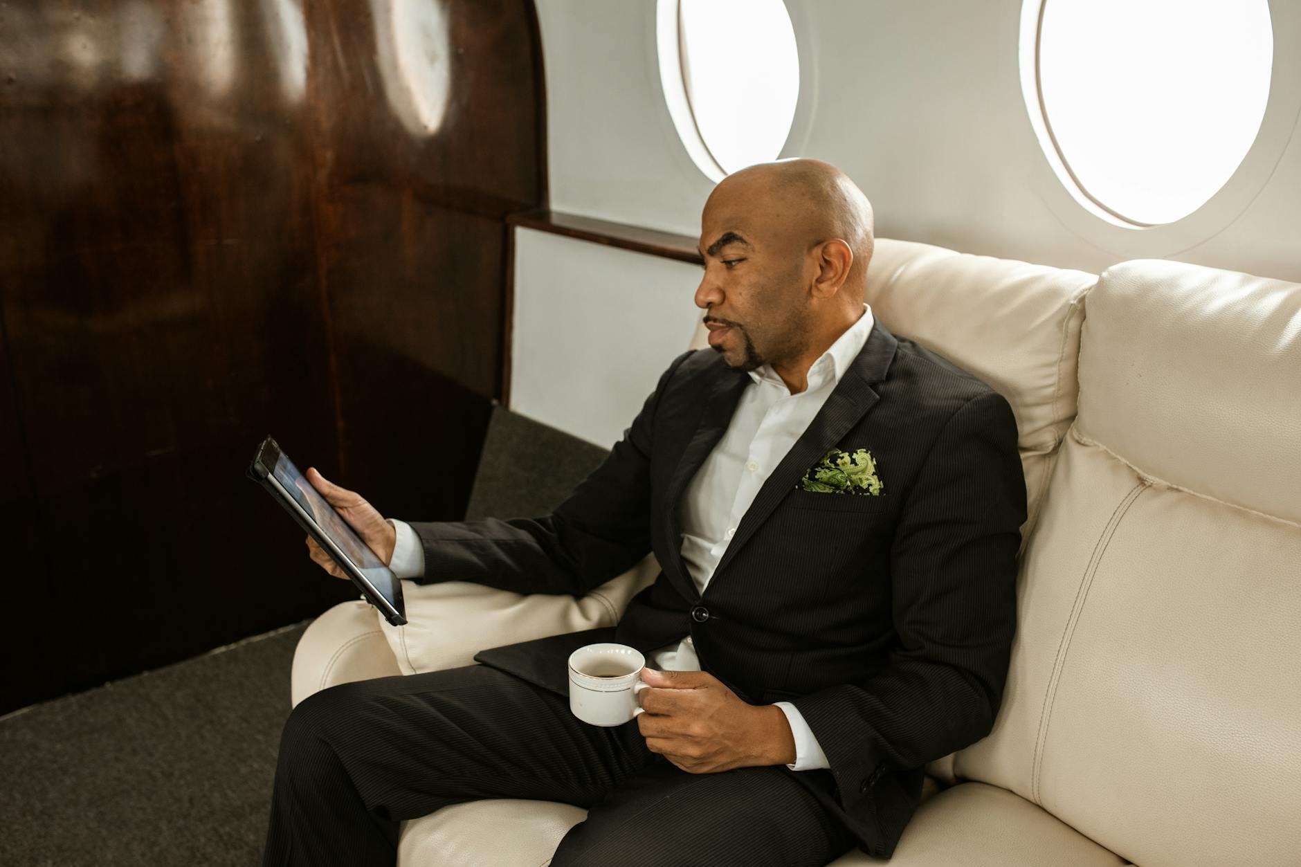 Businessman in formal attire holding tablet and coffee, sitting in private jet.