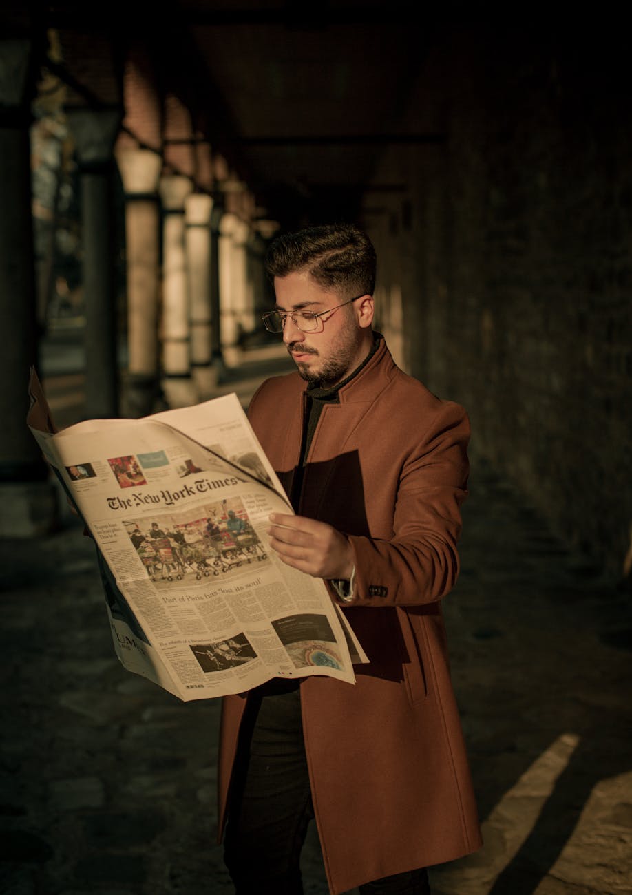 Stylish man reading The New York Times in a city alleyway during sunset.