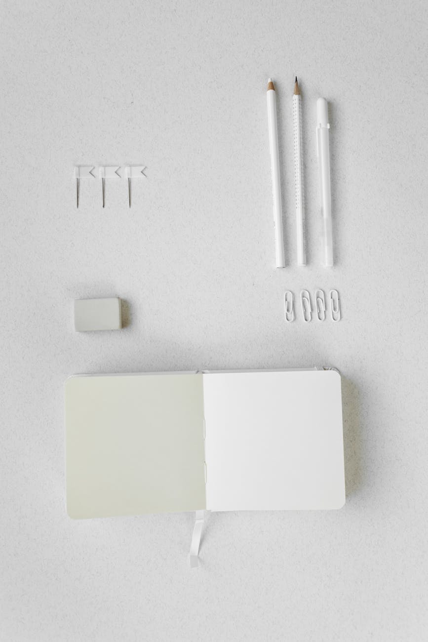 Flat lay of notebook, pencils, eraser, and paper clips on a white surface, ideal for stationery themes.