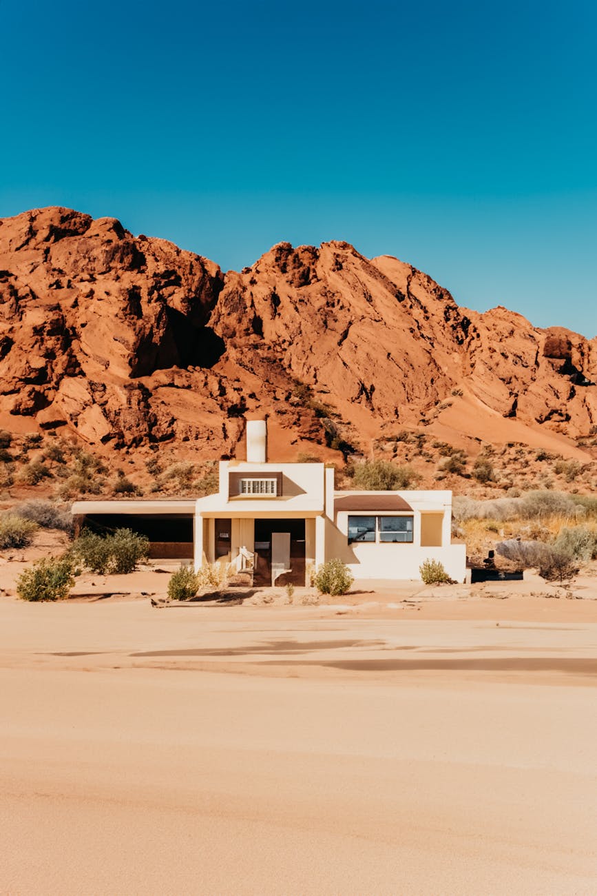 A solitary house stands amidst the arid desert sands and red rocky formations under a clear blue sky.