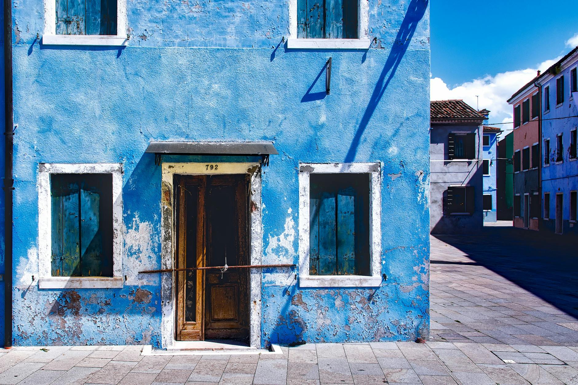 An old, rustic blue building with wooden windows captured in bright daylight on an empty street.