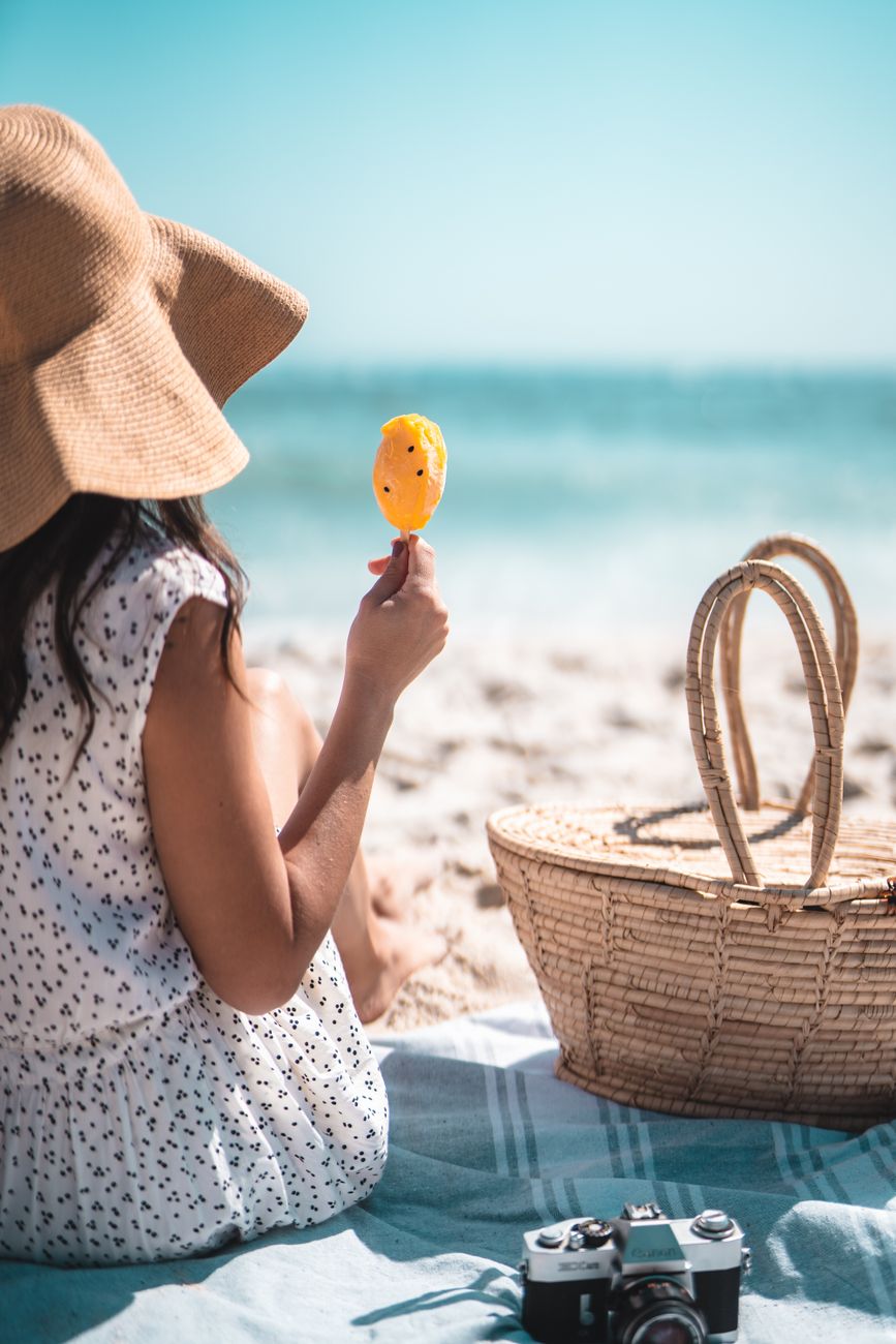 Woman enjoying a summer beach day with a popsicle, basket, and camera.