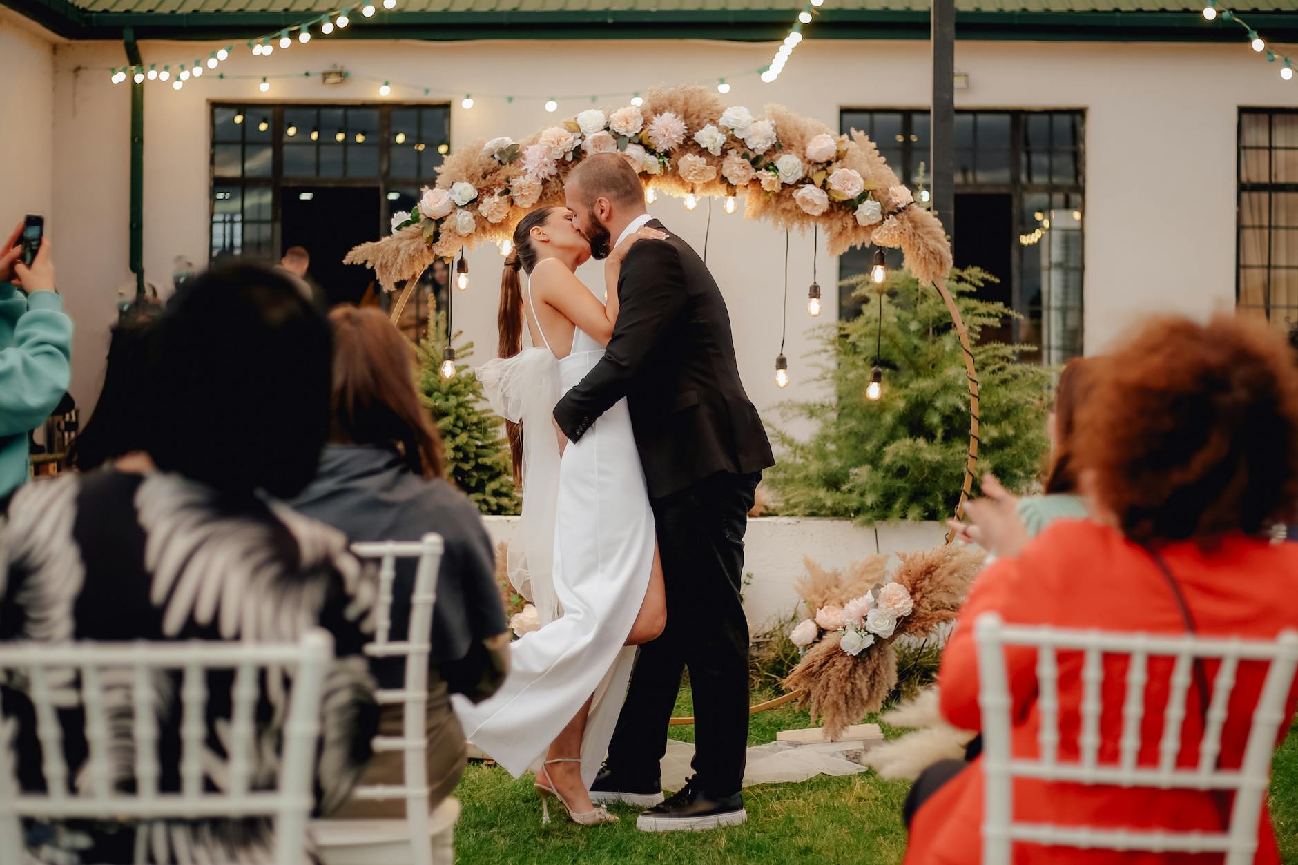 Bride and groom share a kiss under a floral arch during an outdoor wedding ceremony.
