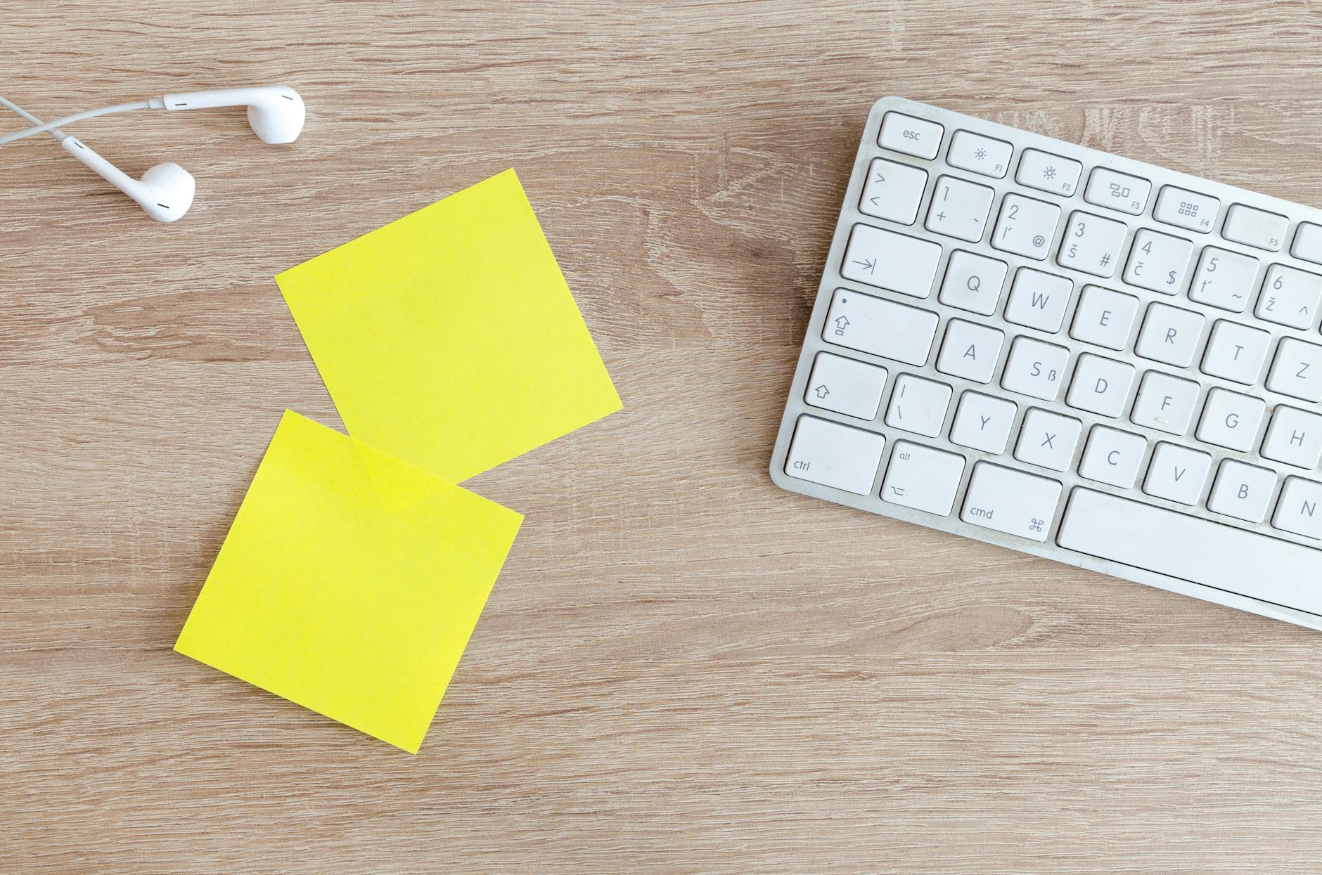 Top view of a workspace with a keyboard, earphones, and yellow sticky notes on a wooden desk.