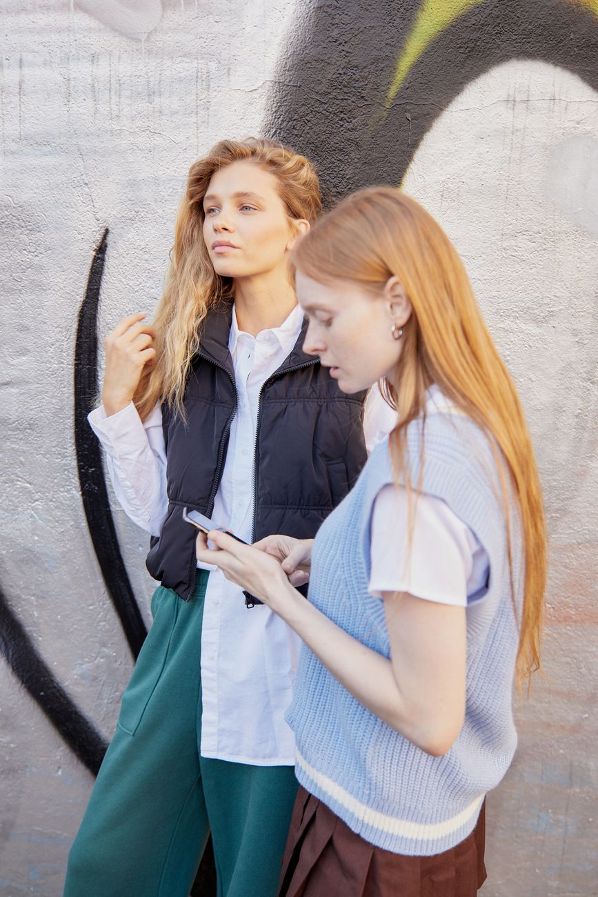 Two fashionable women outdoors engaging with a smartphone, showcasing urban style and friendship.