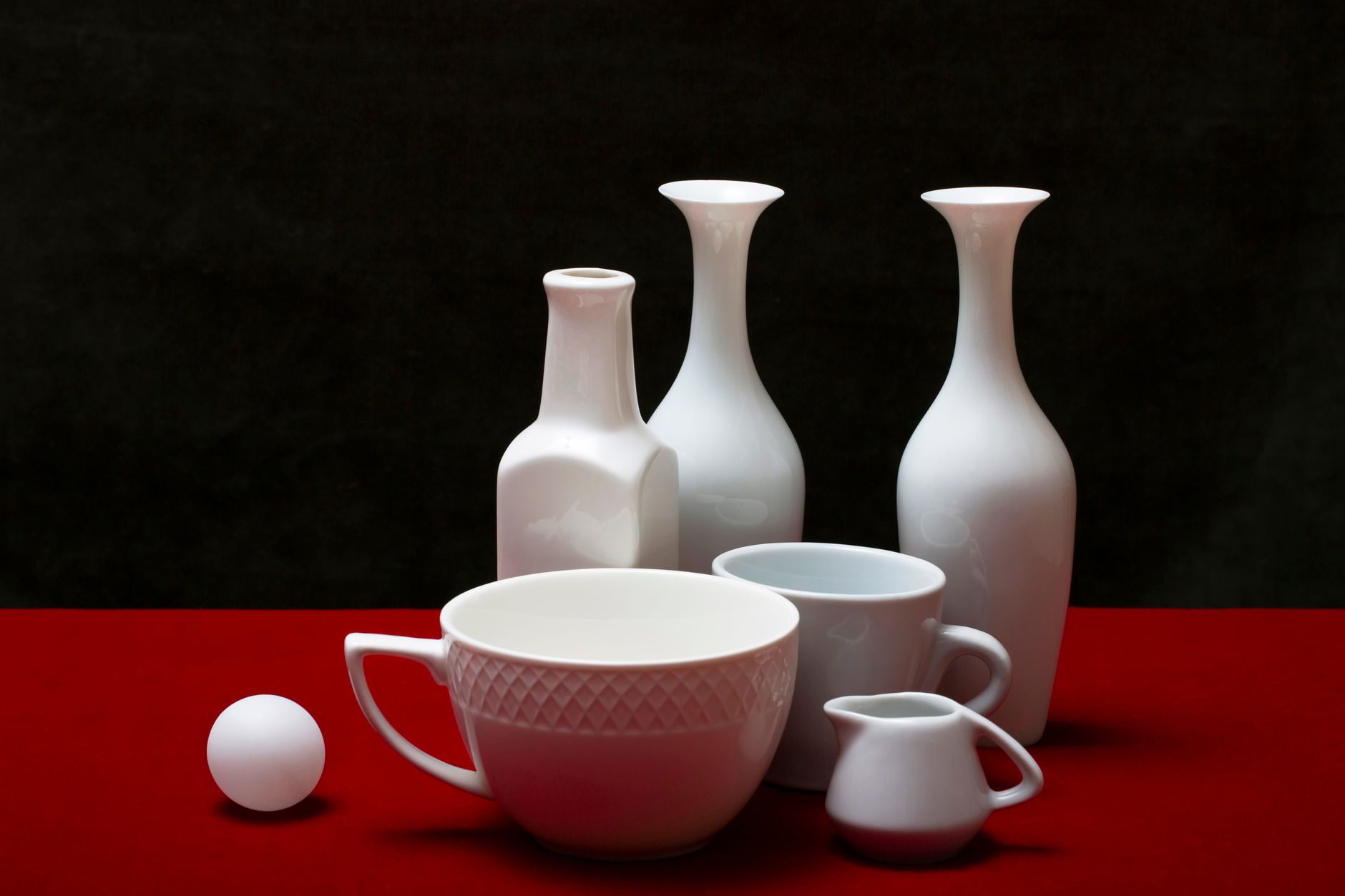 A sophisticated still life showcasing white porcelain vessels on a red surface with a black background.