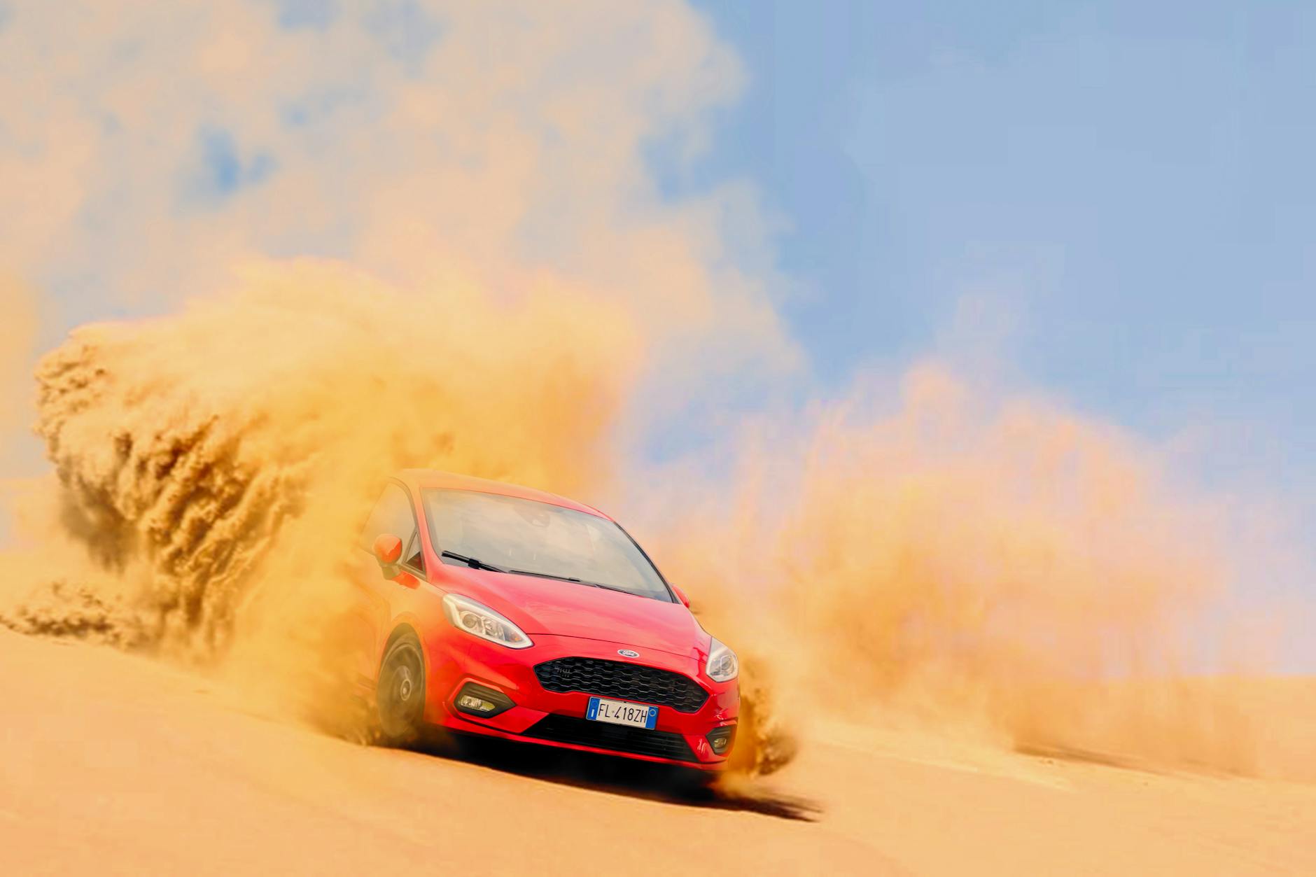 A red car racing at high speed through sandy desert dunes kicking up clouds of dust.