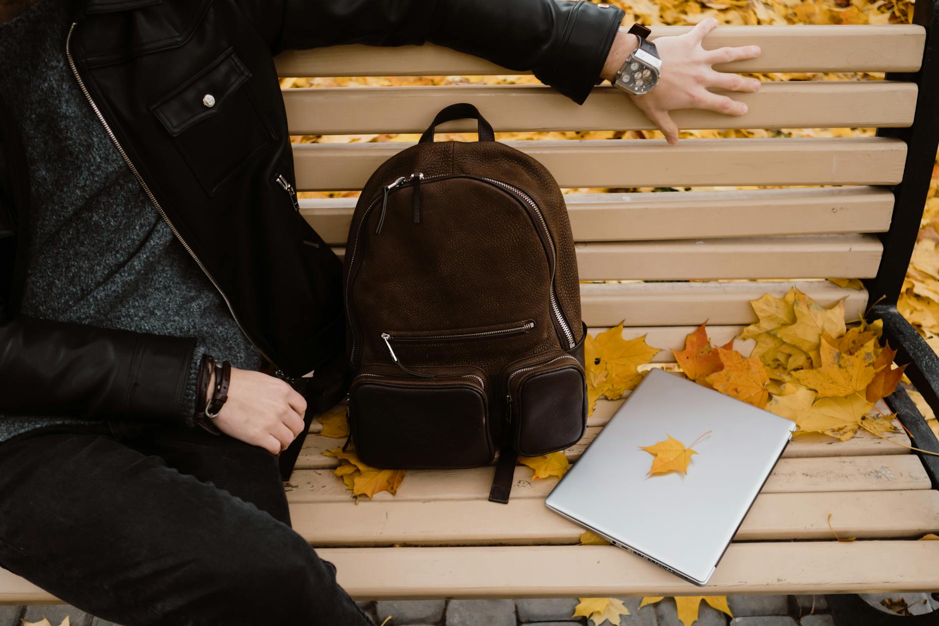 A person sitting on a bench with a backpack and laptop, surrounded by autumn leaves.